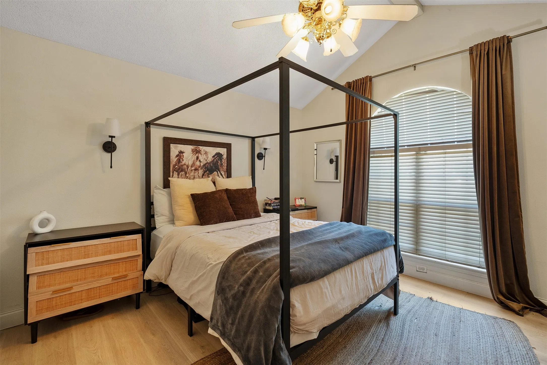 Vaulted ceiling, arched window with blinds, wood-finish flooring, two wall sconces, and a ceiling fan with light fixture