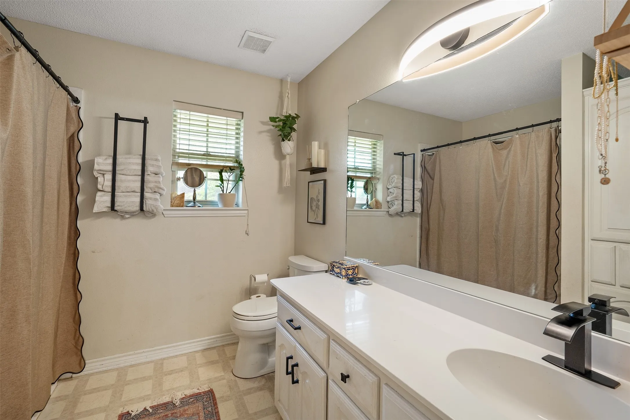 Vanity with an integrated sink and matte black faucet, wall-mounted full-width mirror, windows with blinds, and patterned flooring