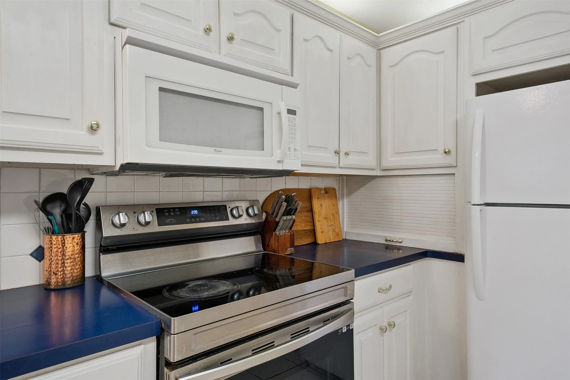 White cabinetry with gold-tone hardware, stainless steel electric range, over-the-range microwave, white tile backsplash, and blue countertops