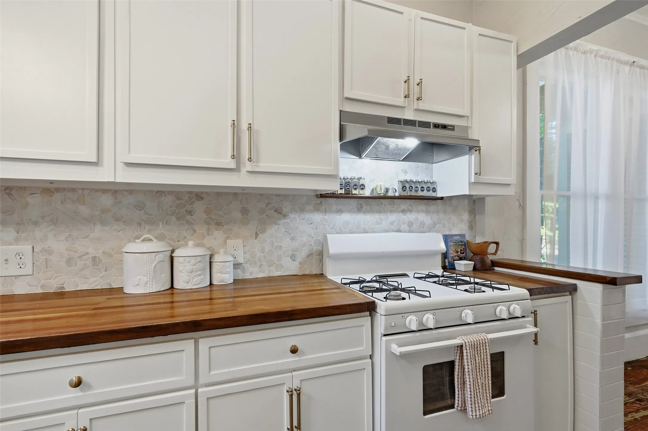White cabinetry with gold-finish hardware, wood-finish countertops, geometric-patterned backsplash, and a white gas range with an overhead exhaust hood