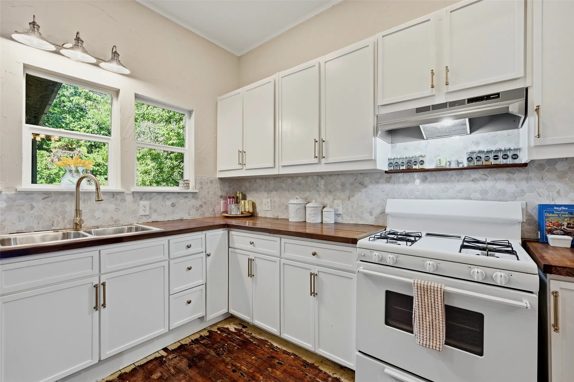 White cabinetry with gold-tone hardware, wood-finish countertops, geometric tile backsplash, and a double basin stainless steel sink