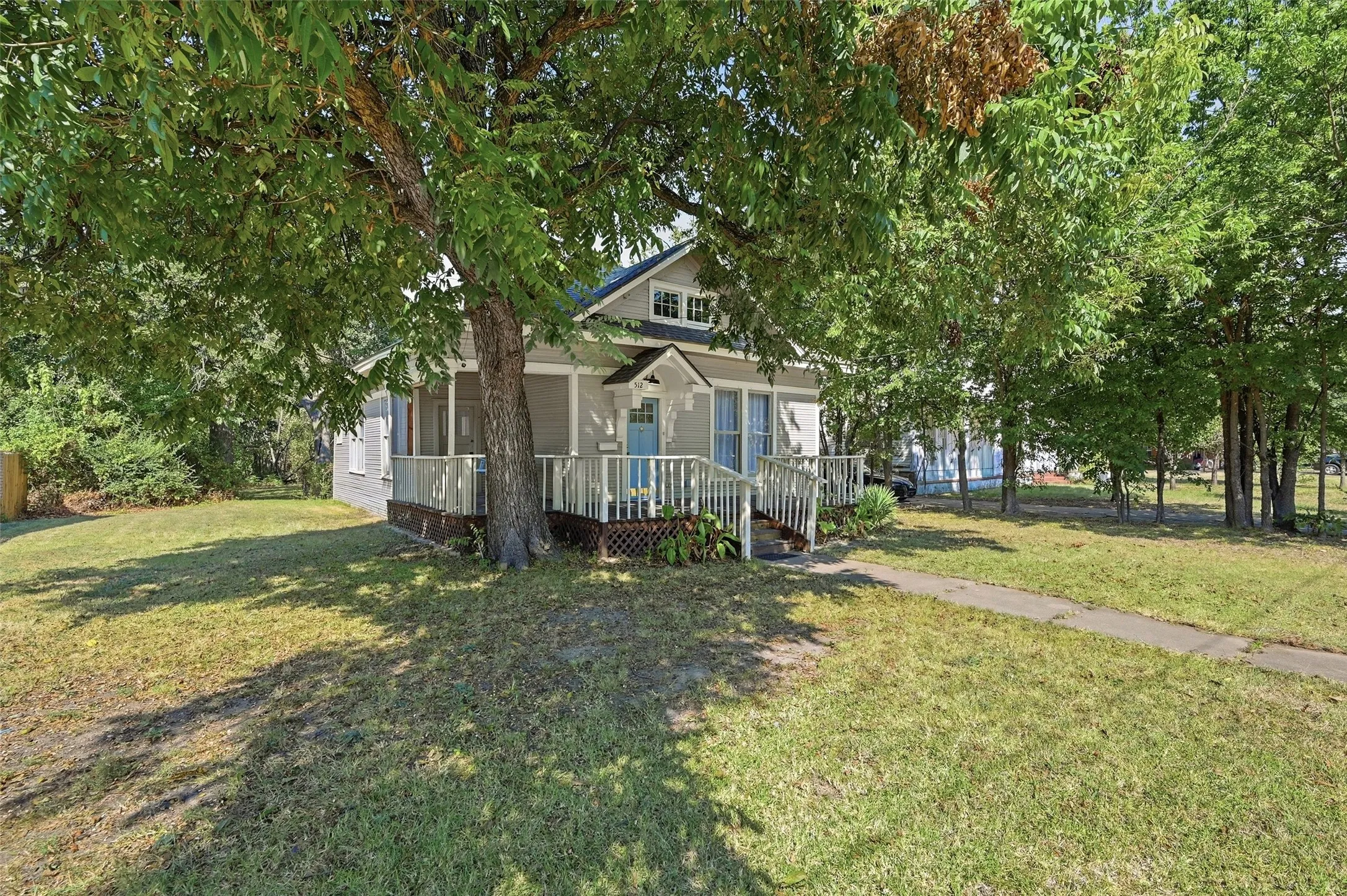 Siding exterior with a covered porch, white railings, a light blue door, and a mature tree providing shade