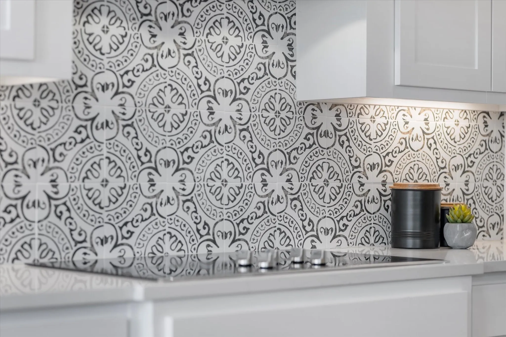 Patterned tile backsplash, white upper cabinetry, built-in cooktop, light-toned countertop, and recessed under-cabinet lighting