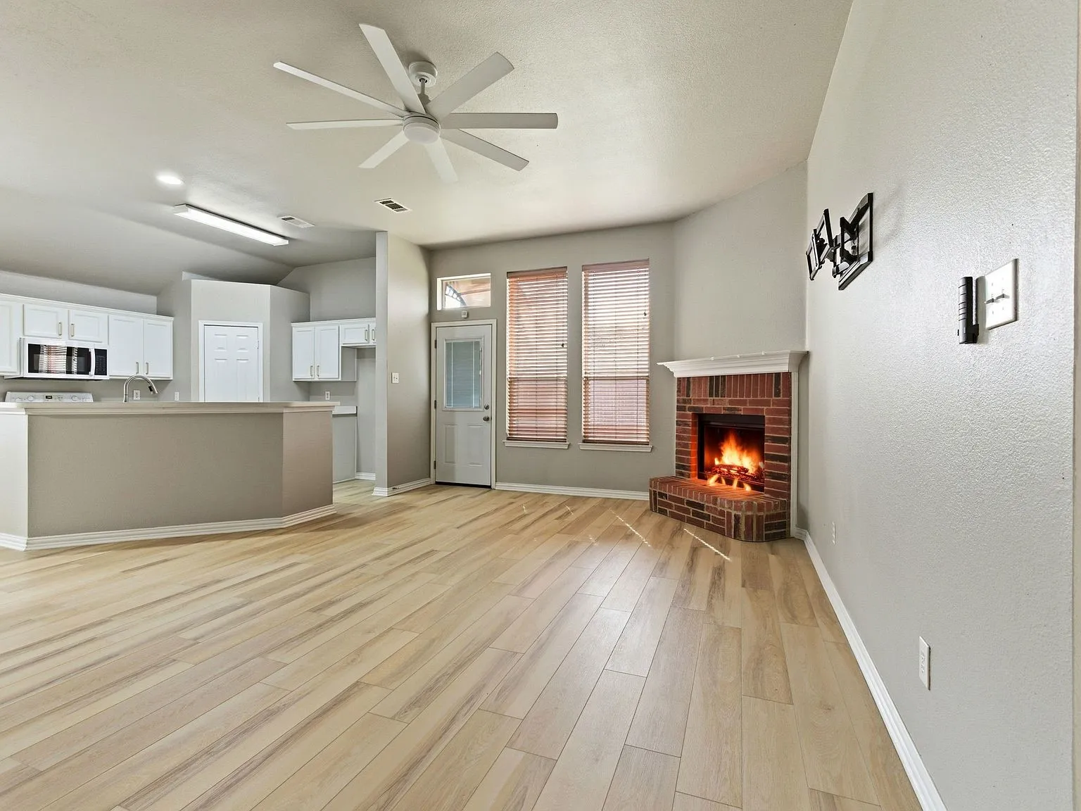 Wood-finish flooring, brick fireplace with mantel, white ceiling fan, multiple windows with blinds, and white kitchen cabinetry