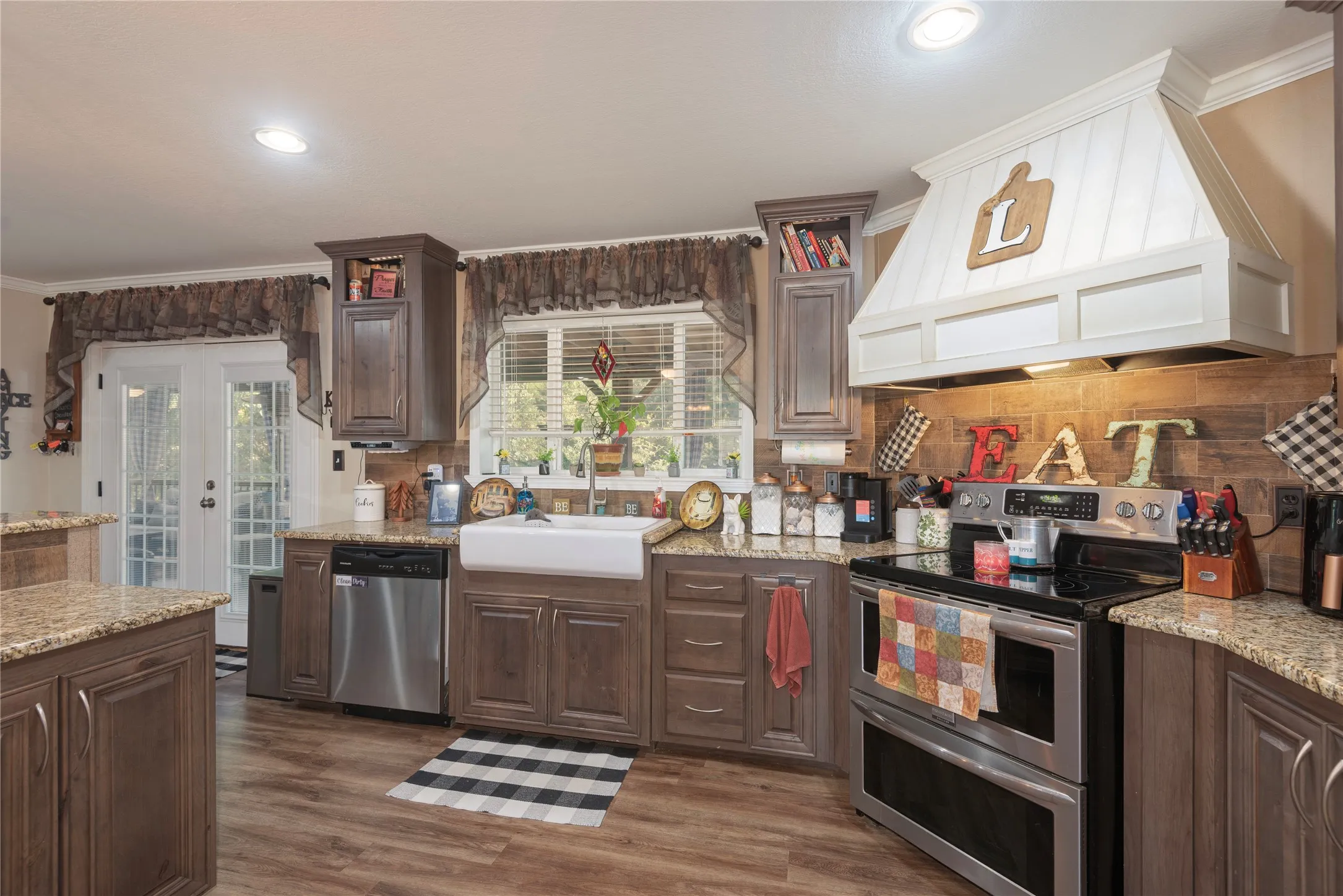 Kitchen featuring wood-finish flooring, dark wood cabinetry, light granite-style countertops, a white farmhouse-style sink, and stainless steel appliances