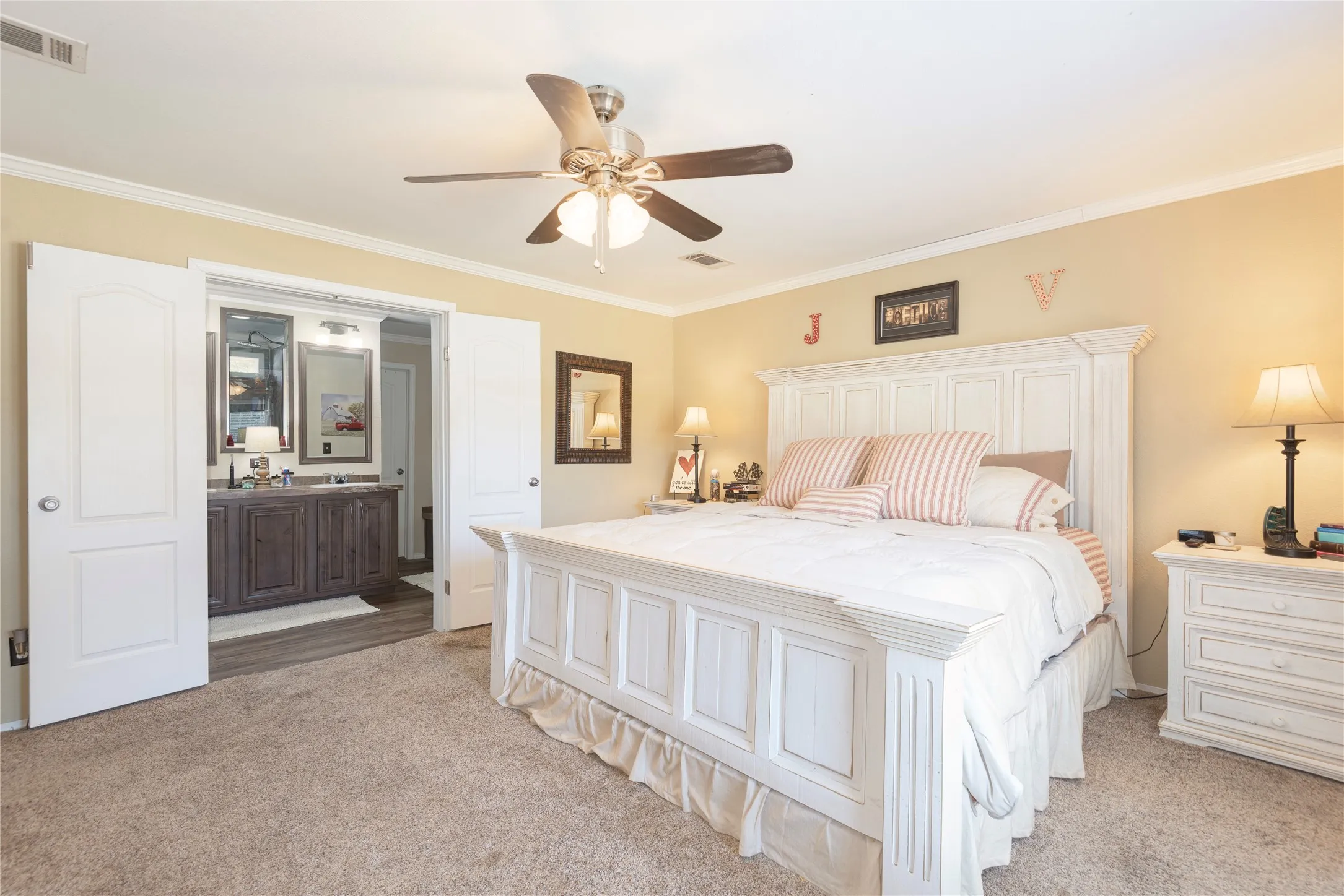 Carpeted bedroom with a ceiling fan, crown molding, and double doors opening to a bathroom featuring a double vanity with an integrated sink and wood-finish flooring