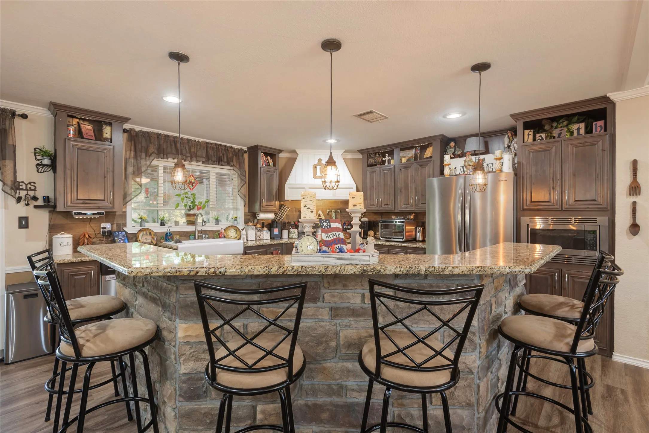 Expansive kitchen island with a stone finish and granite countertop, featuring pendant lighting, wood-finish cabinetry, and stainless steel appliances