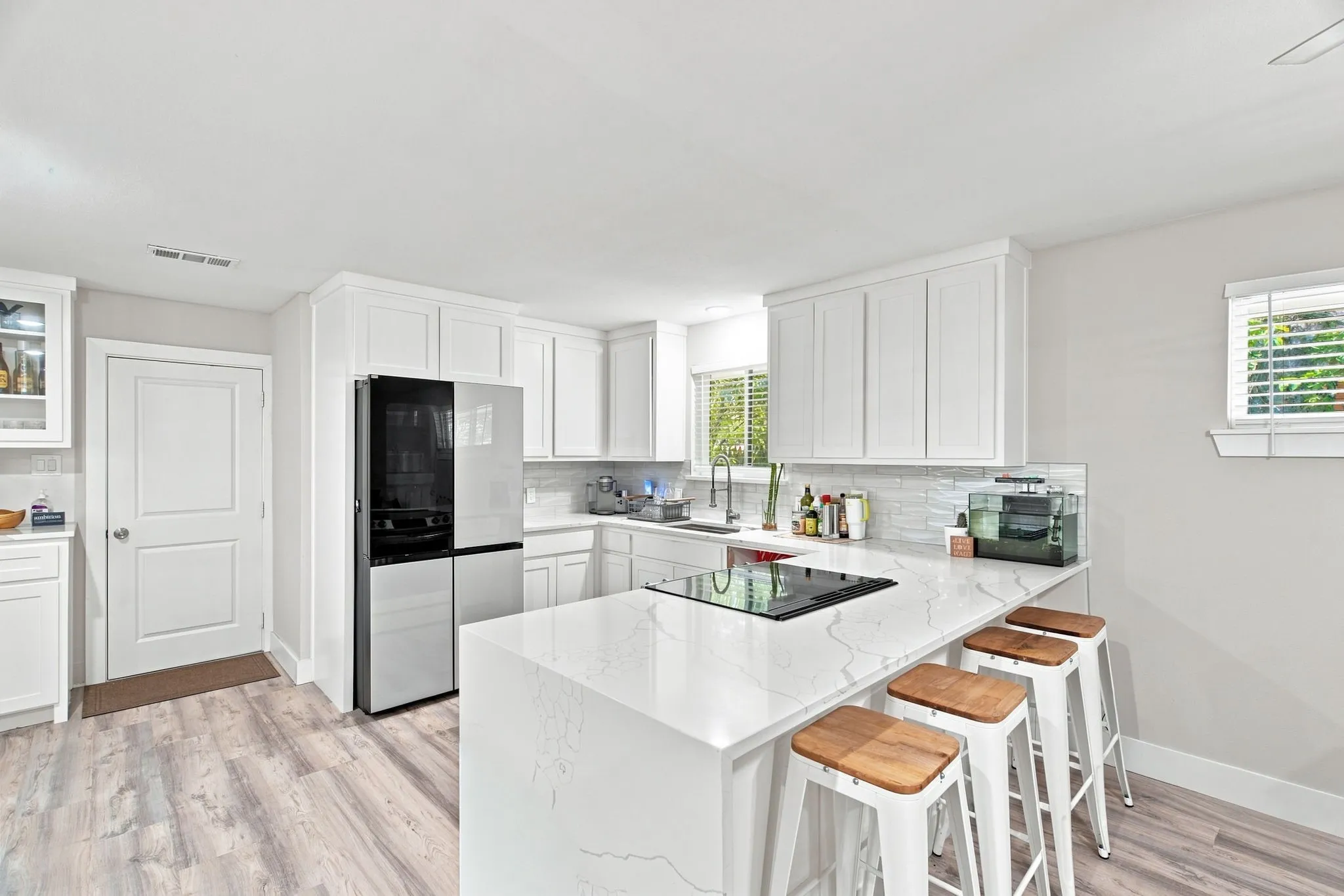 White shaker cabinetry, light wood-finish flooring, built-in cooktop, stainless steel refrigerator, and light countertops