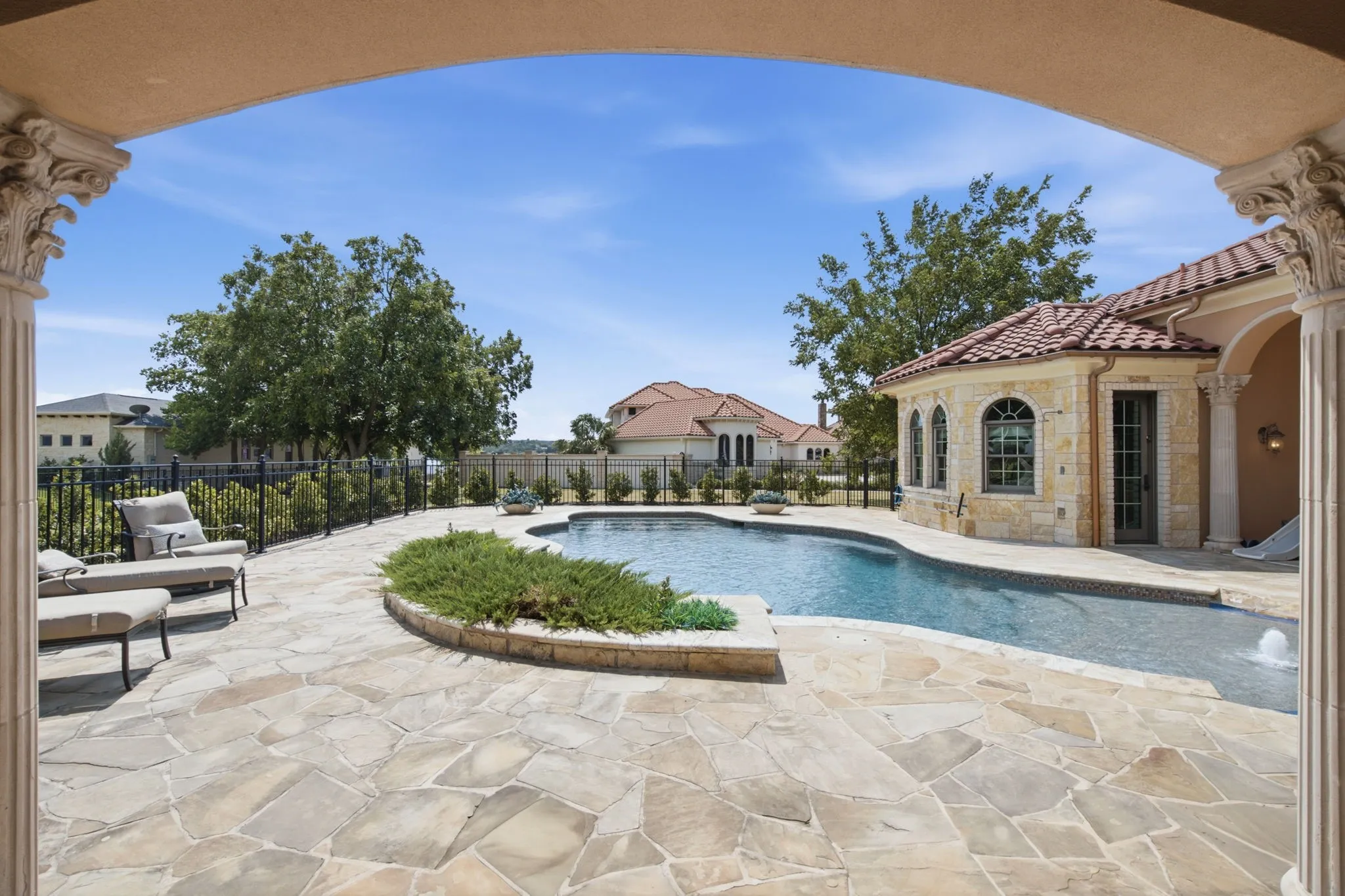 Expansive flagstone patio with a curved swimming pool, surrounded by a wrought-iron fence and mature landscaping