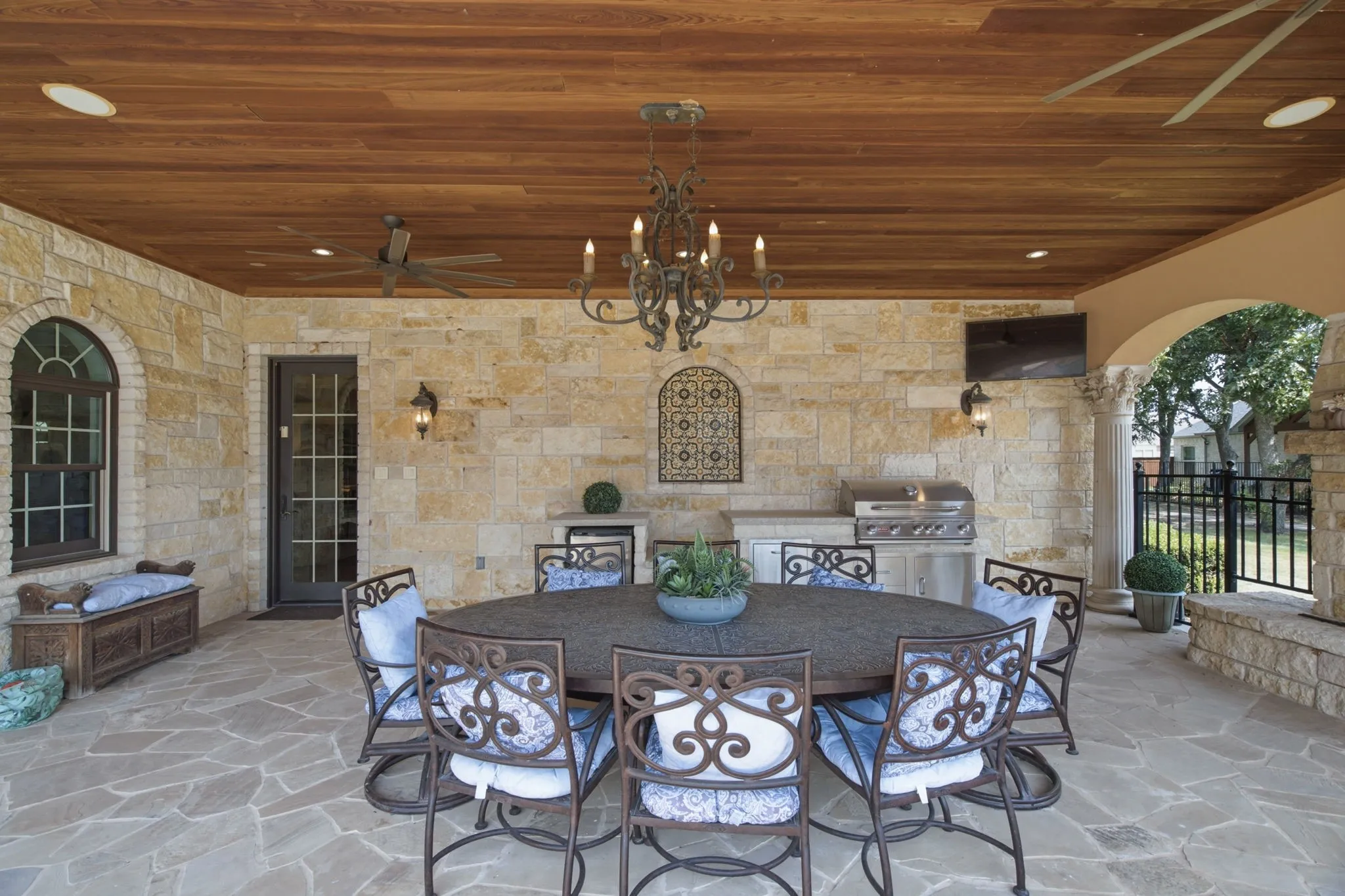Covered outdoor patio featuring a wood-finish ceiling, stone wall cladding, flagstone flooring, built-in outdoor kitchen with grill, and an arched entryway