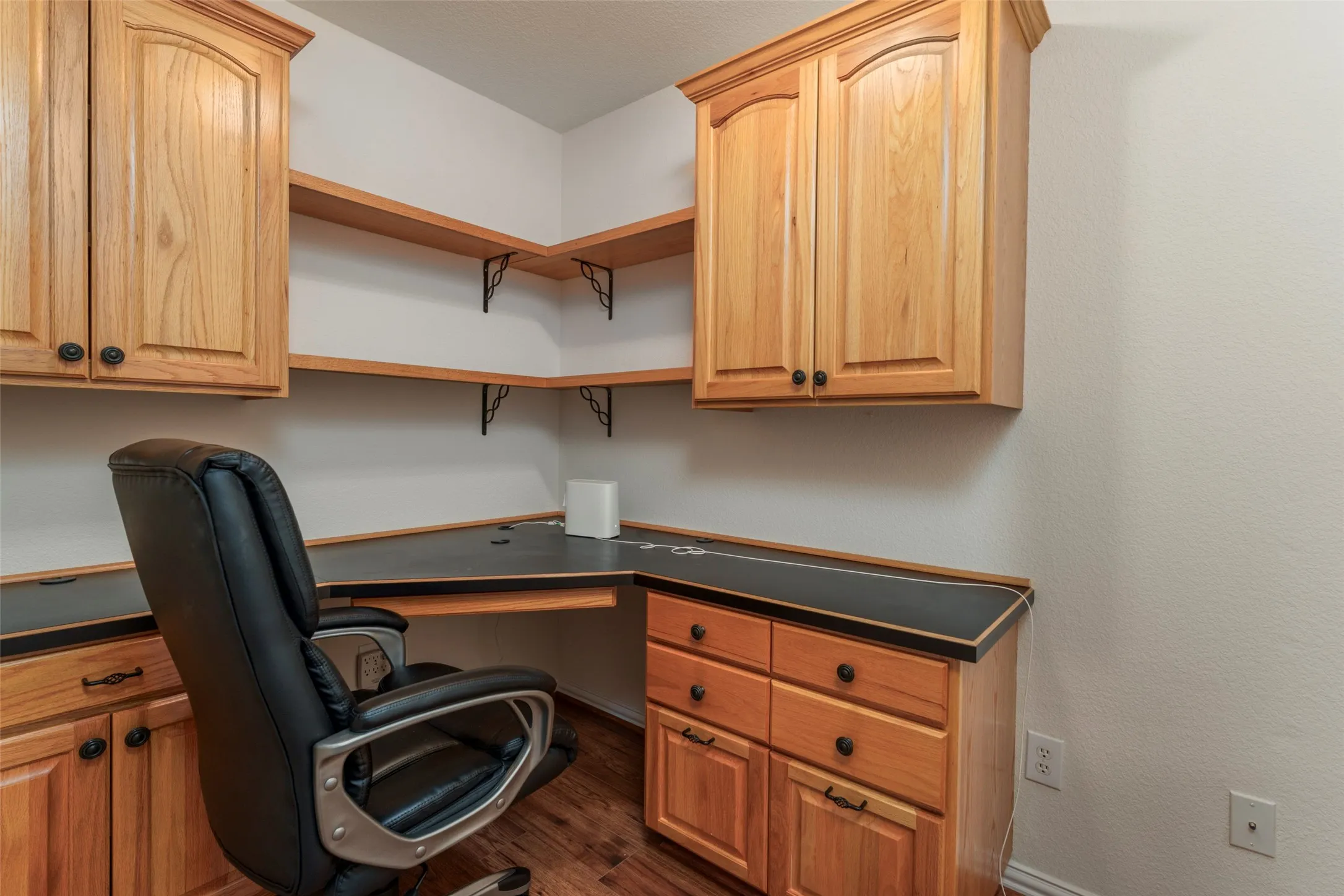 Built-in cabinetry with wood-finish doors and dark hardware, corner desk with a dark countertop, integrated shelving, and wood-finish flooring