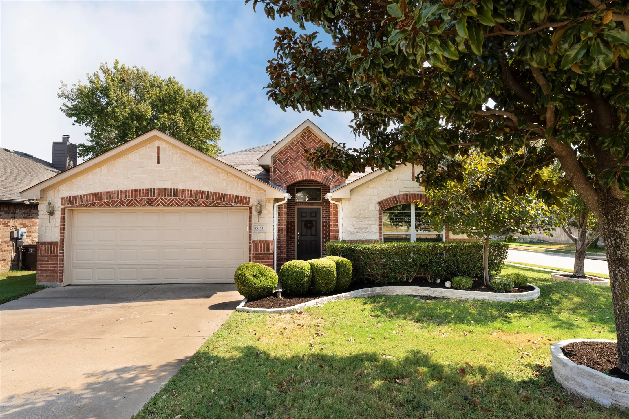 Brick and stone exterior with arched accents, attached two-car garage, mature front yard tree, and landscaped garden beds