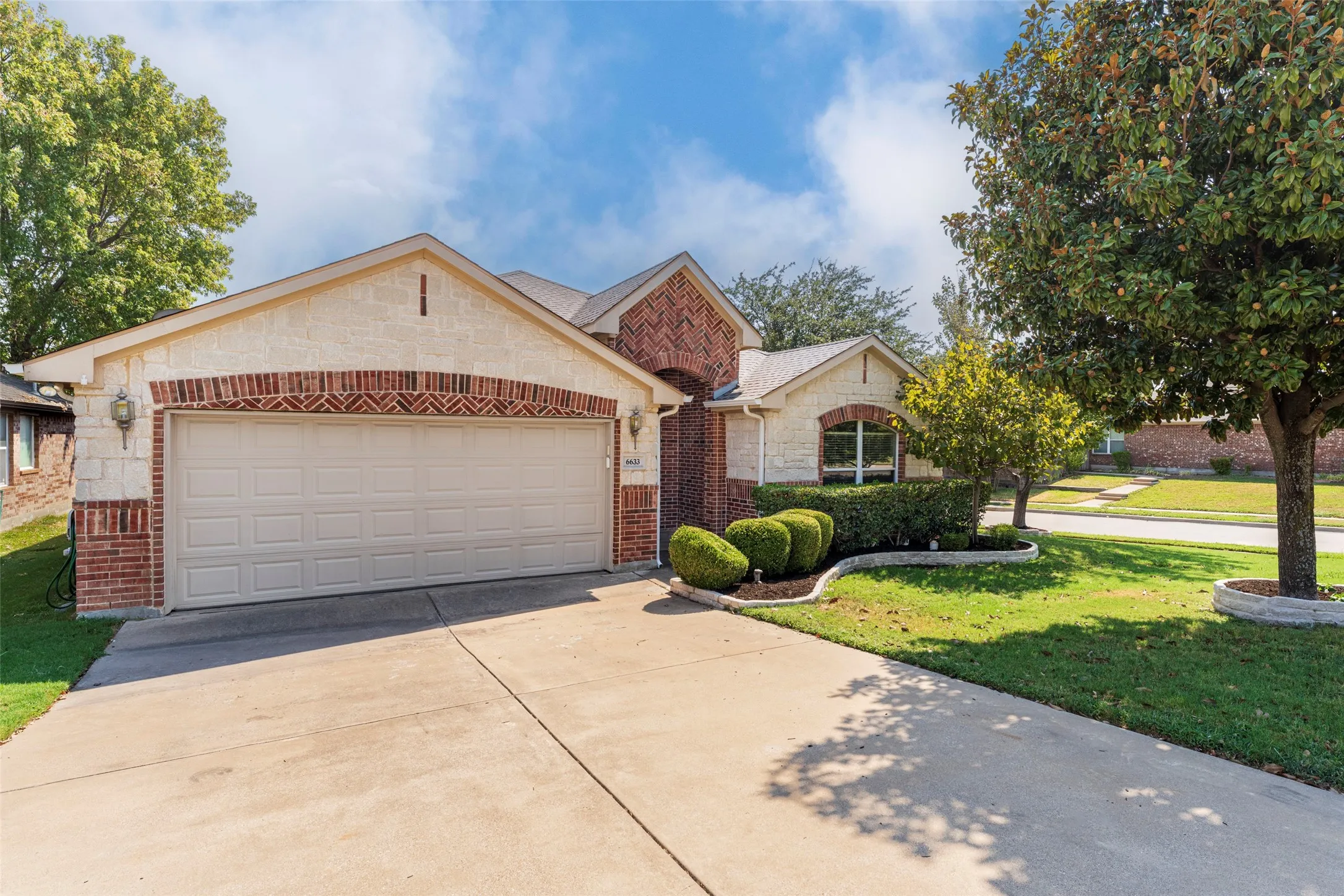 Brick and stone façade with an arched entryway, attached two-car garage, concrete driveway, and established front landscaping