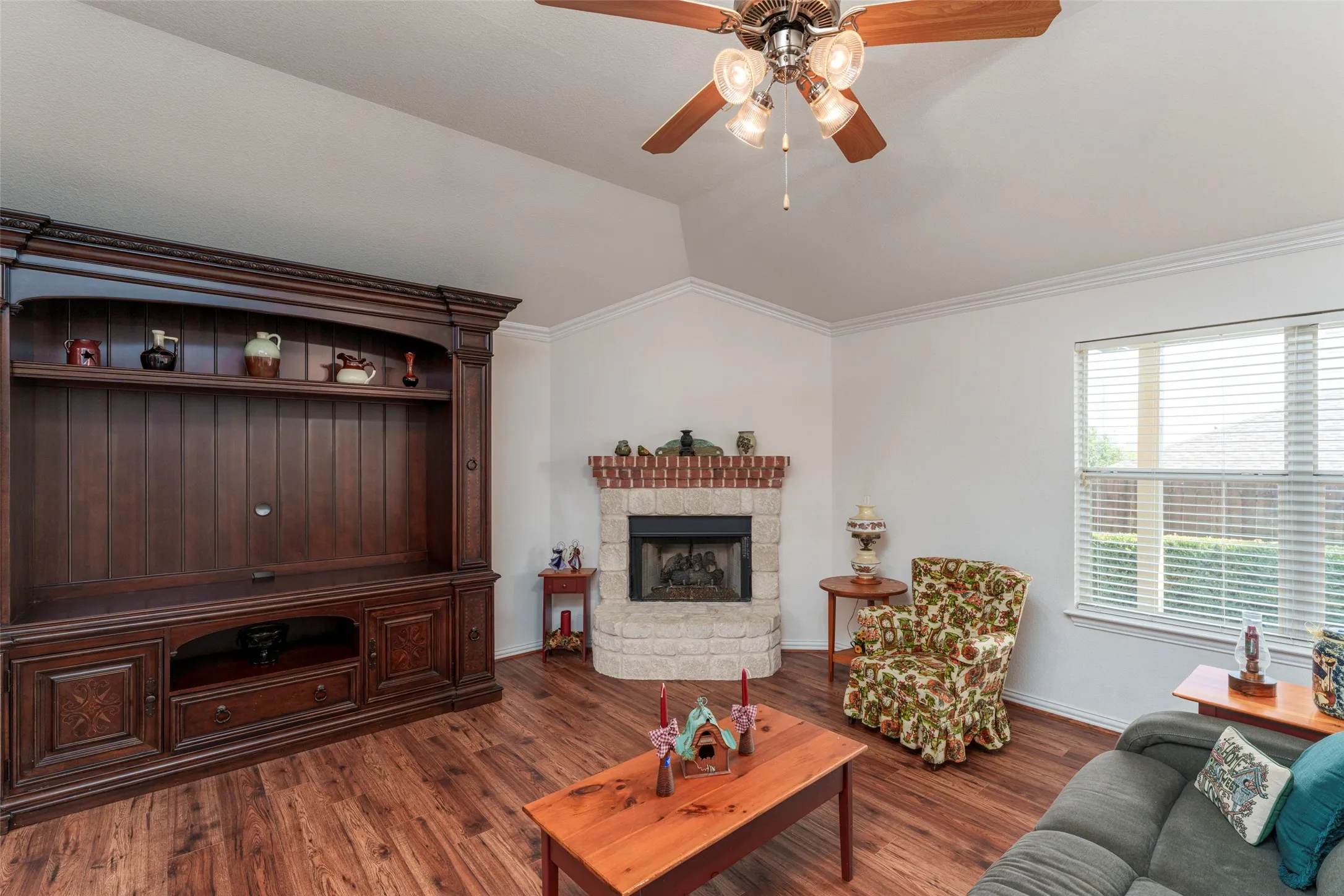 Vaulted ceiling with a ceiling fan light fixture, wood-finish flooring, fireplace with a stone surround and brick mantel, and a window with blinds