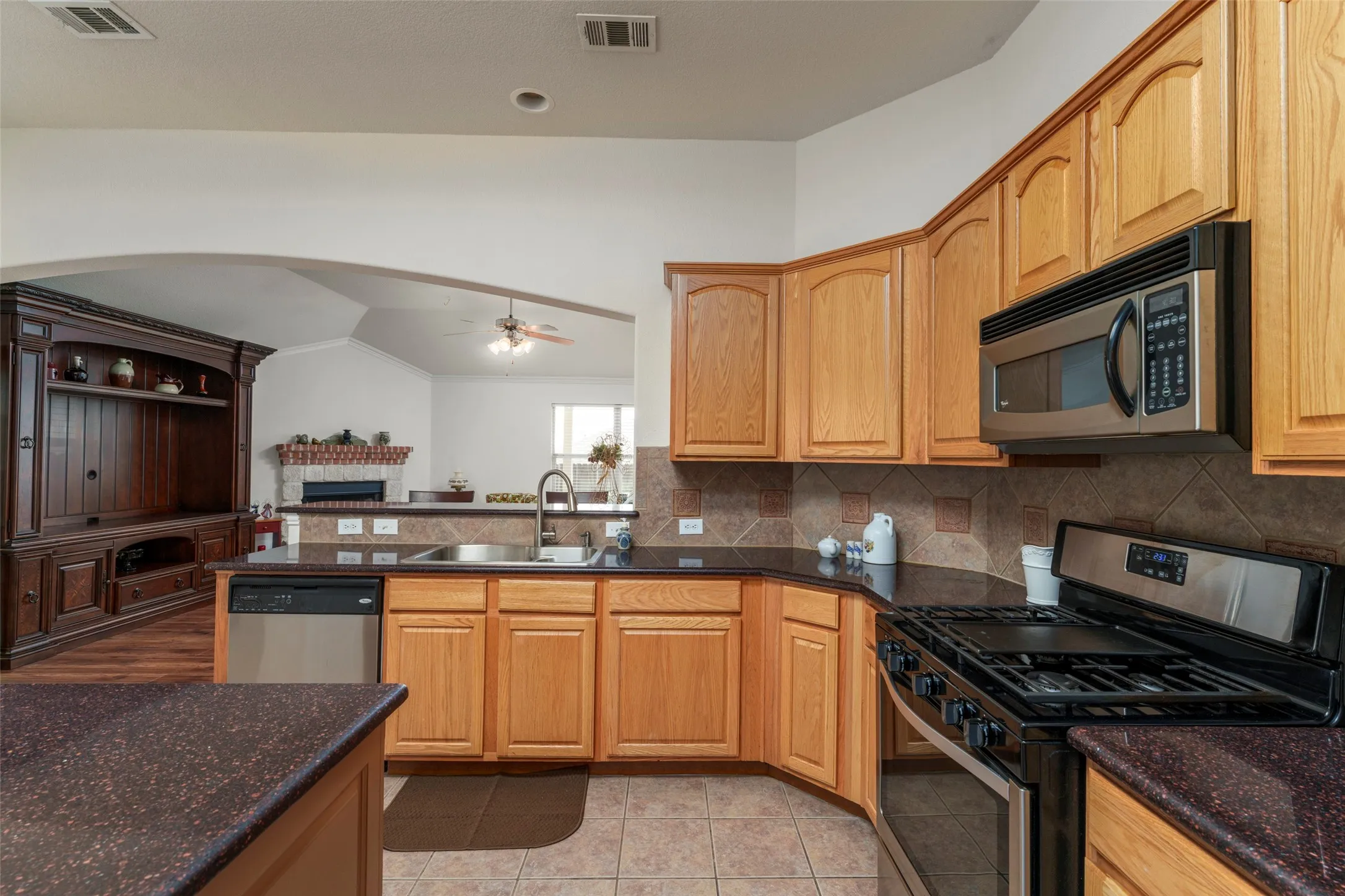 Chef's kitchen featuring warm wood cabinetry, stainless steel gas range and microwave, dark stone countertops, and tile backsplash