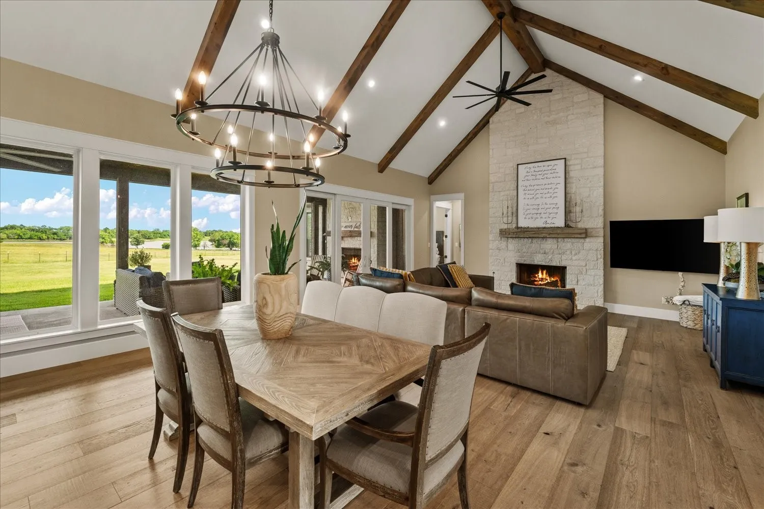 Vaulted ceiling with exposed wood beams, stone fireplace, wood-finish flooring, and large windows with outdoor access