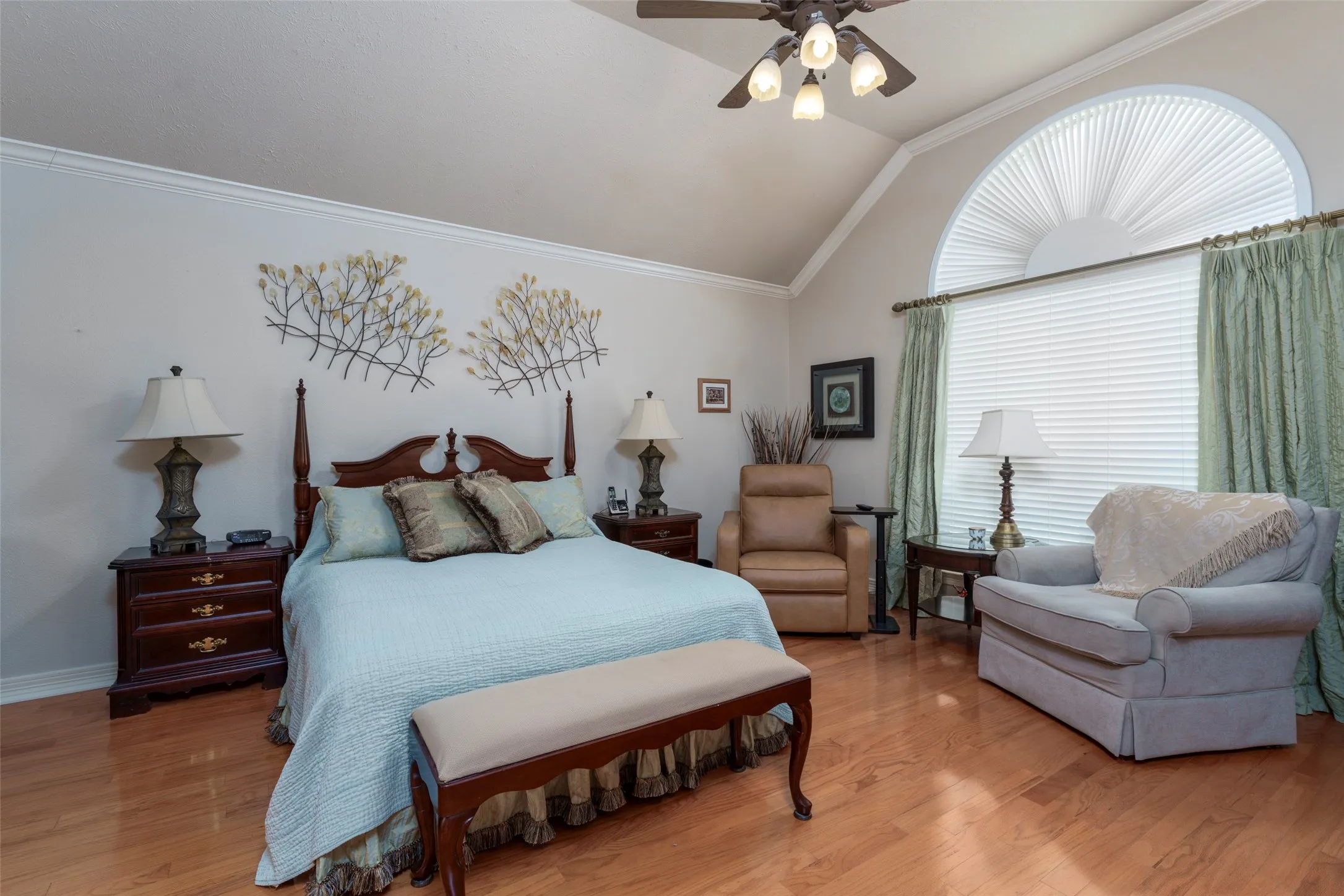 Spacious primary bedroom with hardwood flooring, ceiling fan with integrated lighting, and arched transom window with plantation shutters