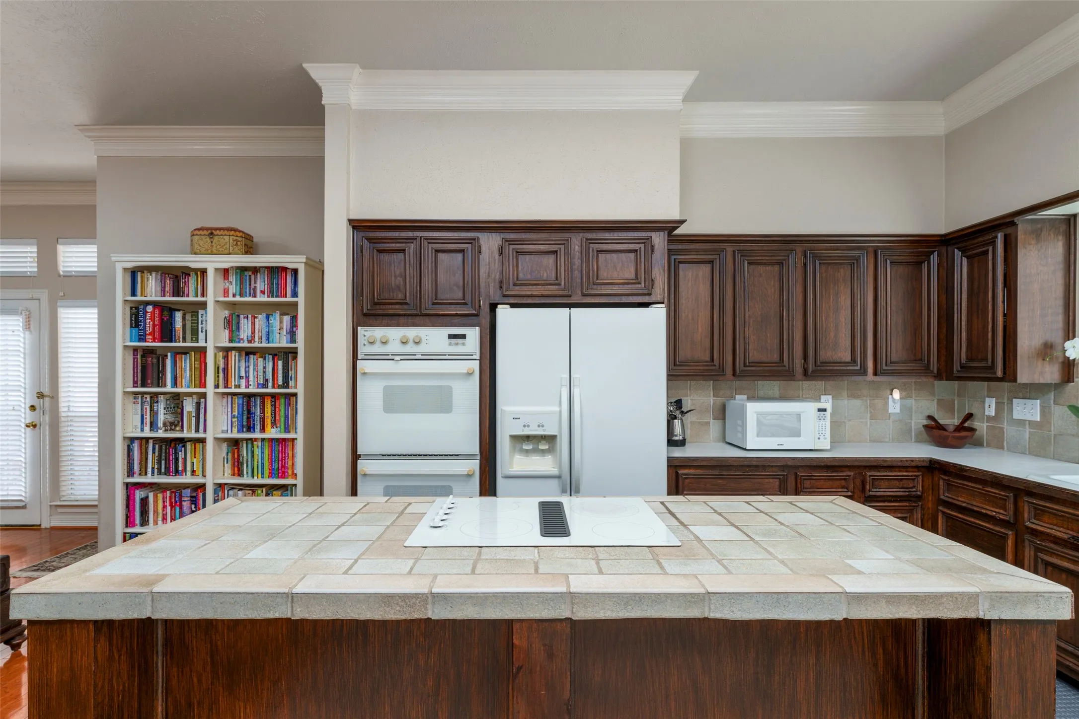 Kitchen island with tile countertop and integrated cooktop, complemented by dark wood-finish cabinetry, built-in double ovens, and a tile backsplash