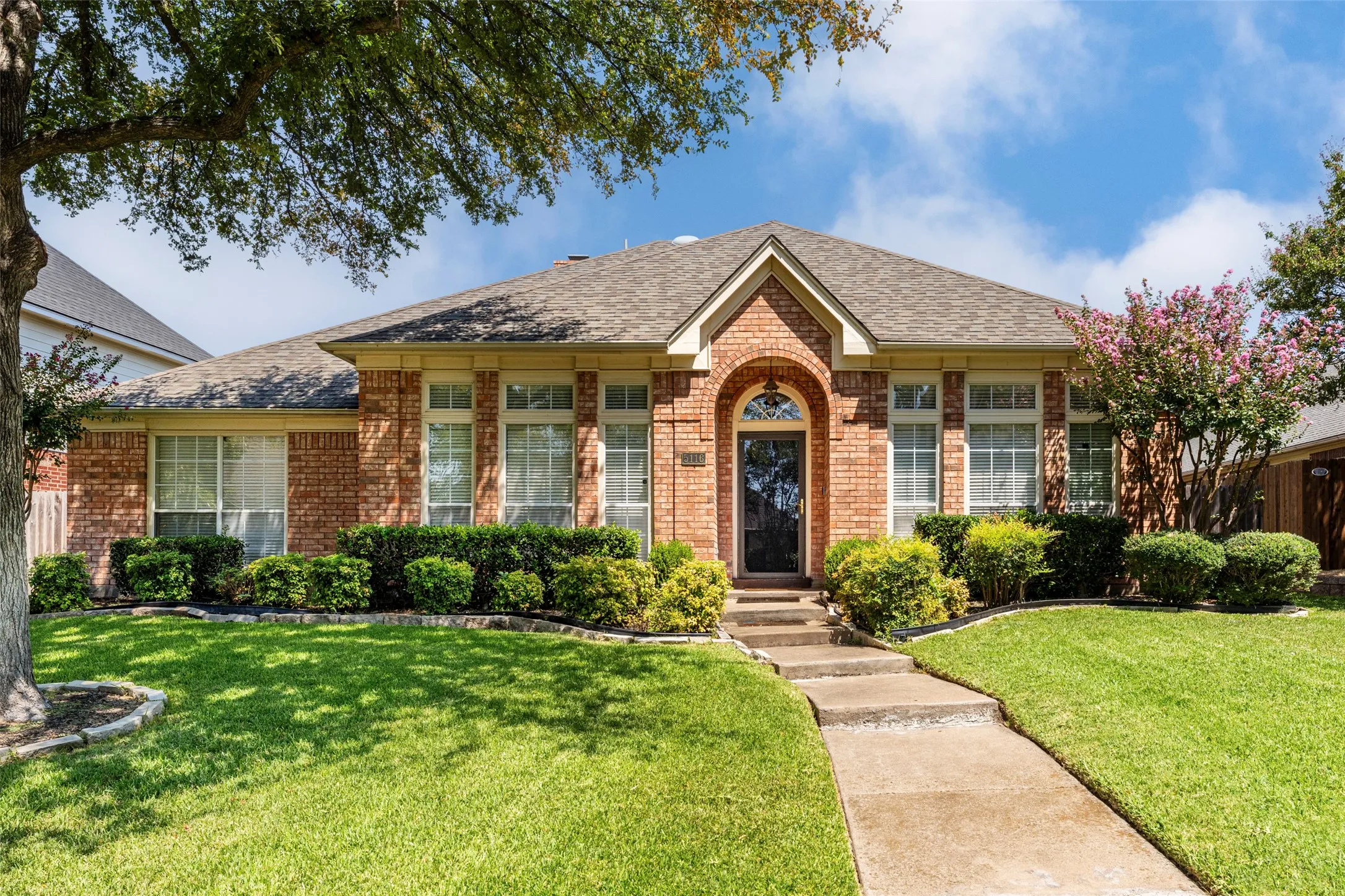 Brick exterior with arched entryway, front lawn with mature trees, and established shrubbery