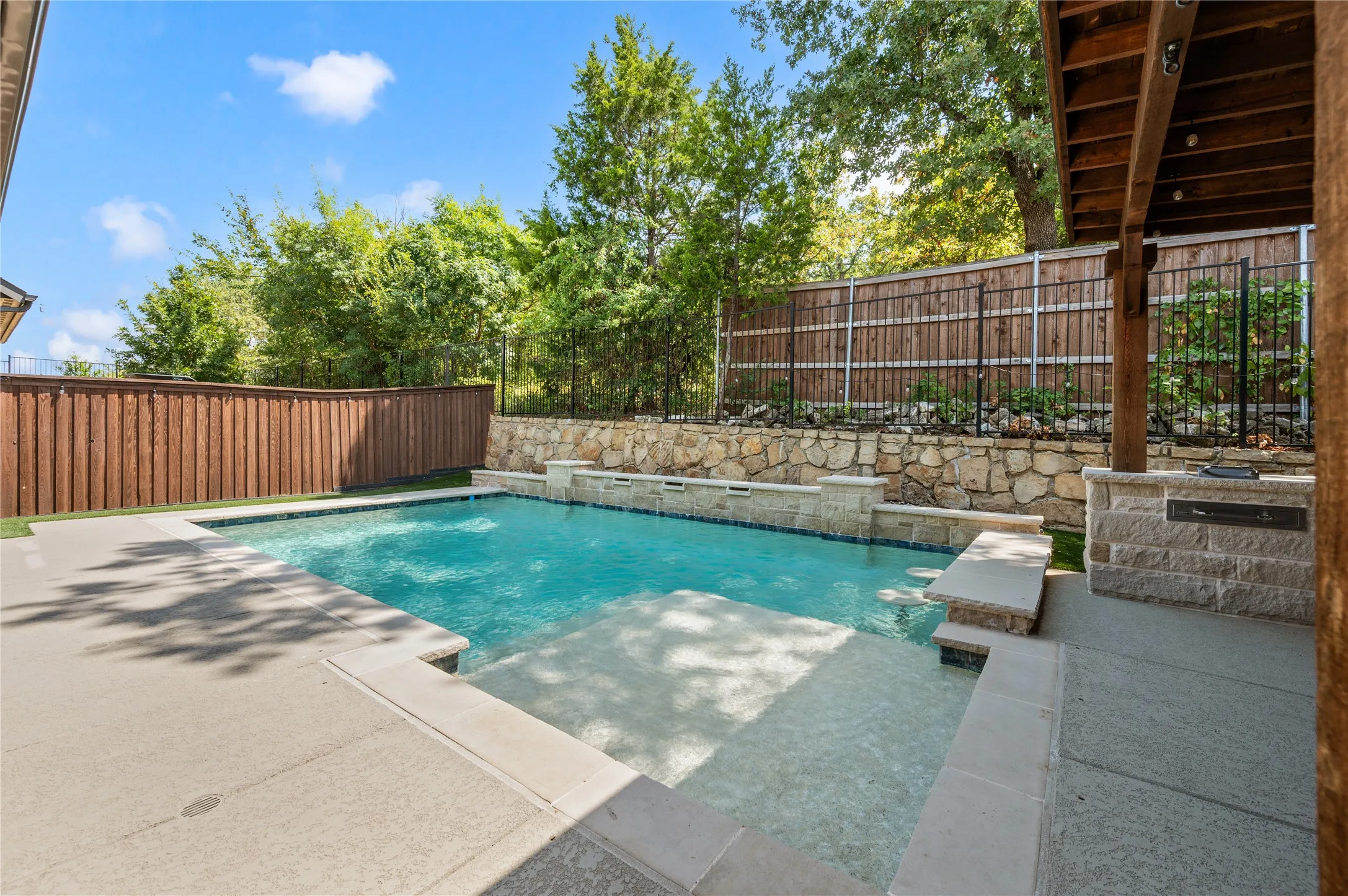 Swimming pool with integrated sun shelf, stone-clad retaining wall, water features, and a built-in outdoor kitchen area