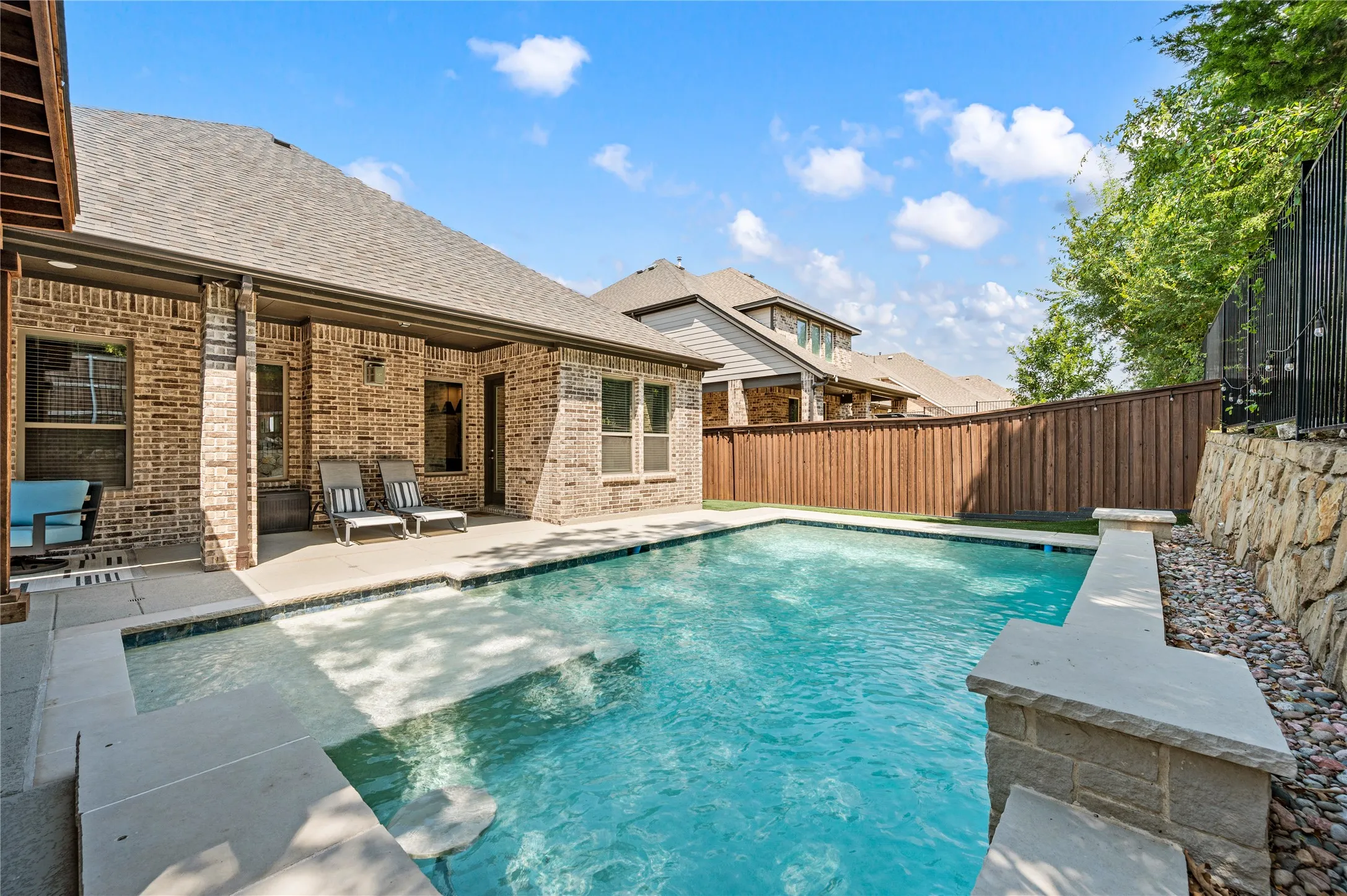 Outdoor swimming pool with integrated tanning ledge, stone retaining wall, brick exterior, and covered patio area