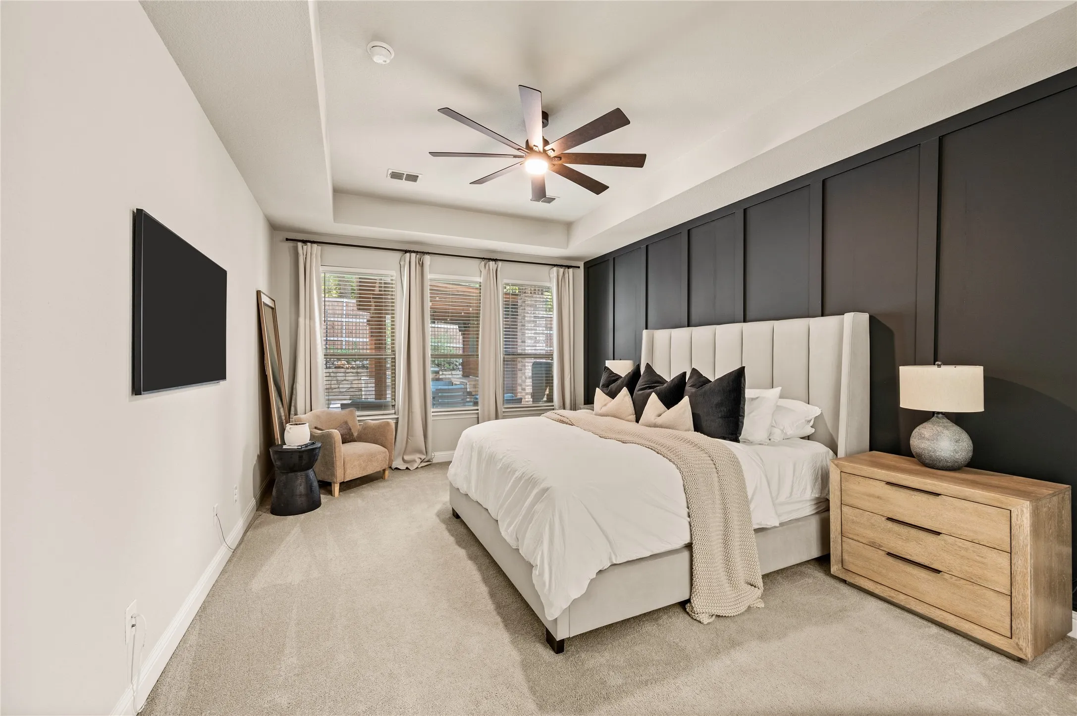 Sophisticated primary bedroom featuring a custom upholstered headboard wall in charcoal with white accent paneling, light wood-finish nightstands with multiple drawers, and a modern ceiling fan
