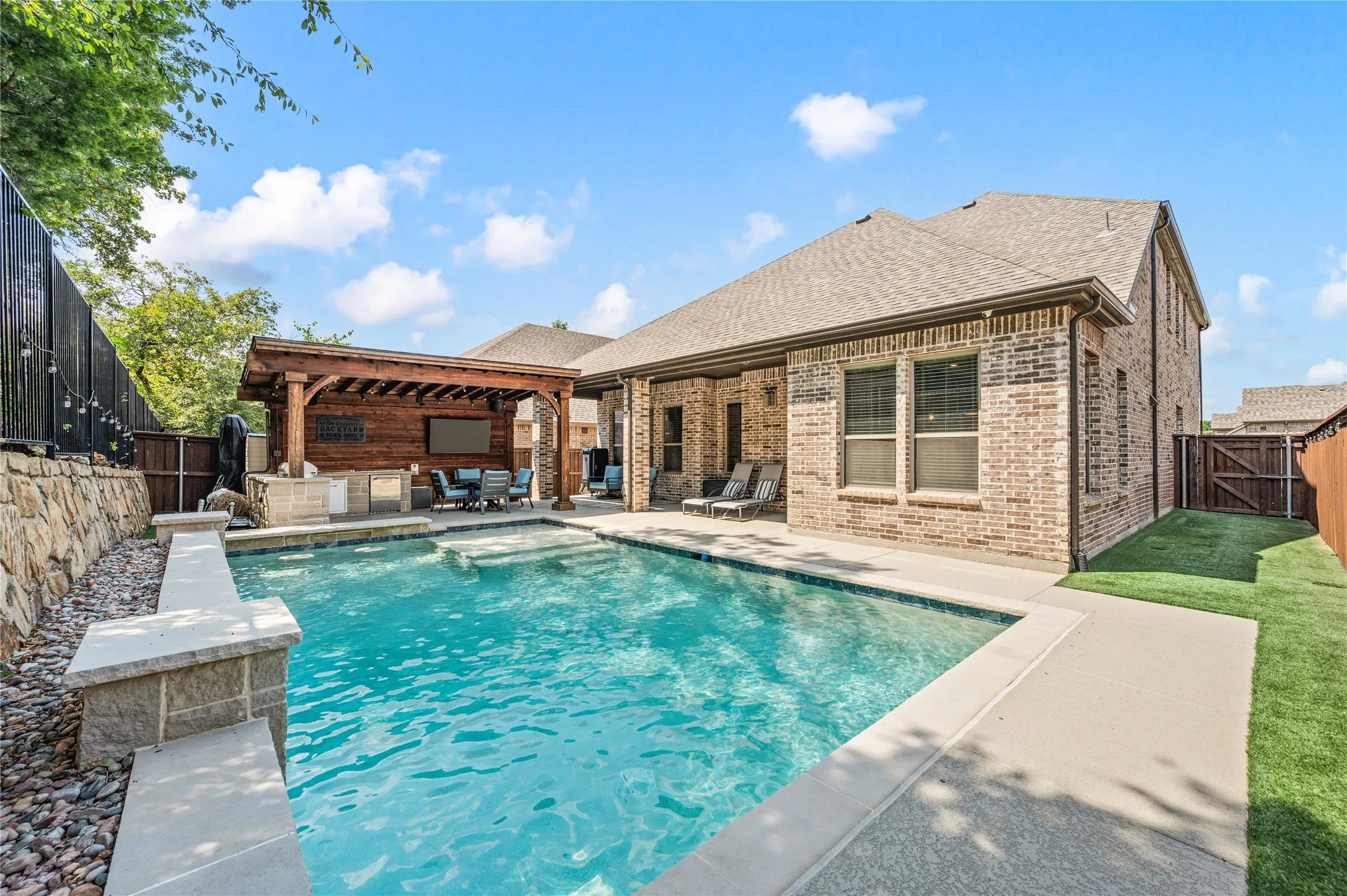 Backyard featuring a rectangular swimming pool with integrated steps, an outdoor kitchen with a wood pergola, brick exterior, and artificial turf