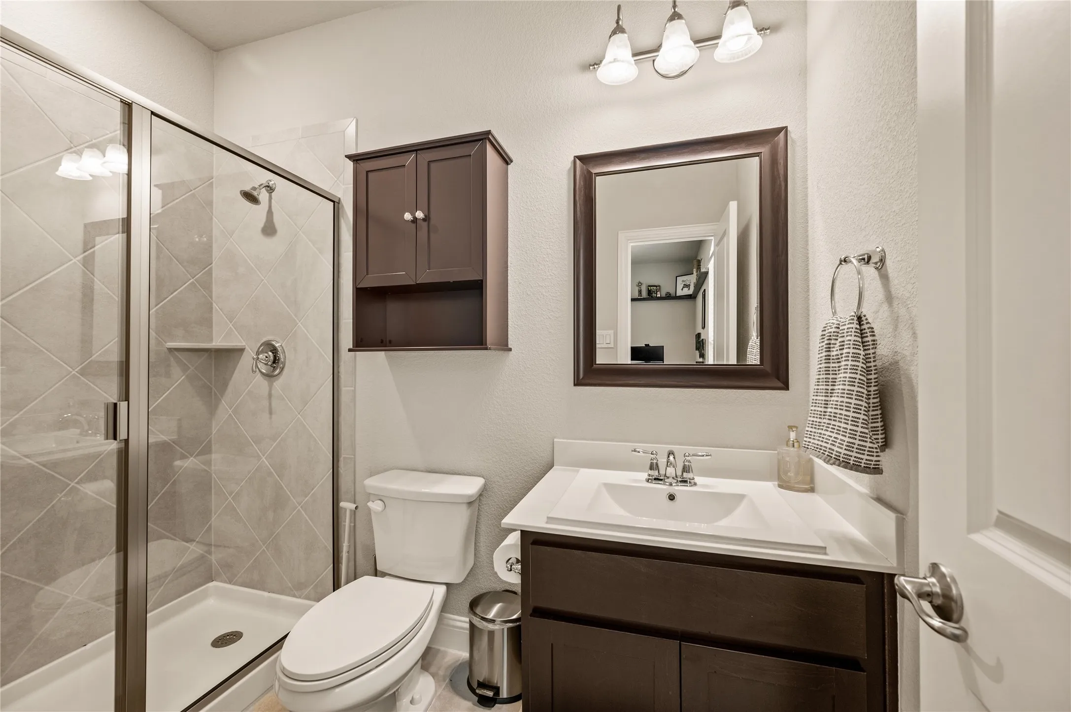 Glass-enclosed shower with diagonal tilework, white vanity with integrated sink, dark wood-finish cabinetry, and a framed mirror