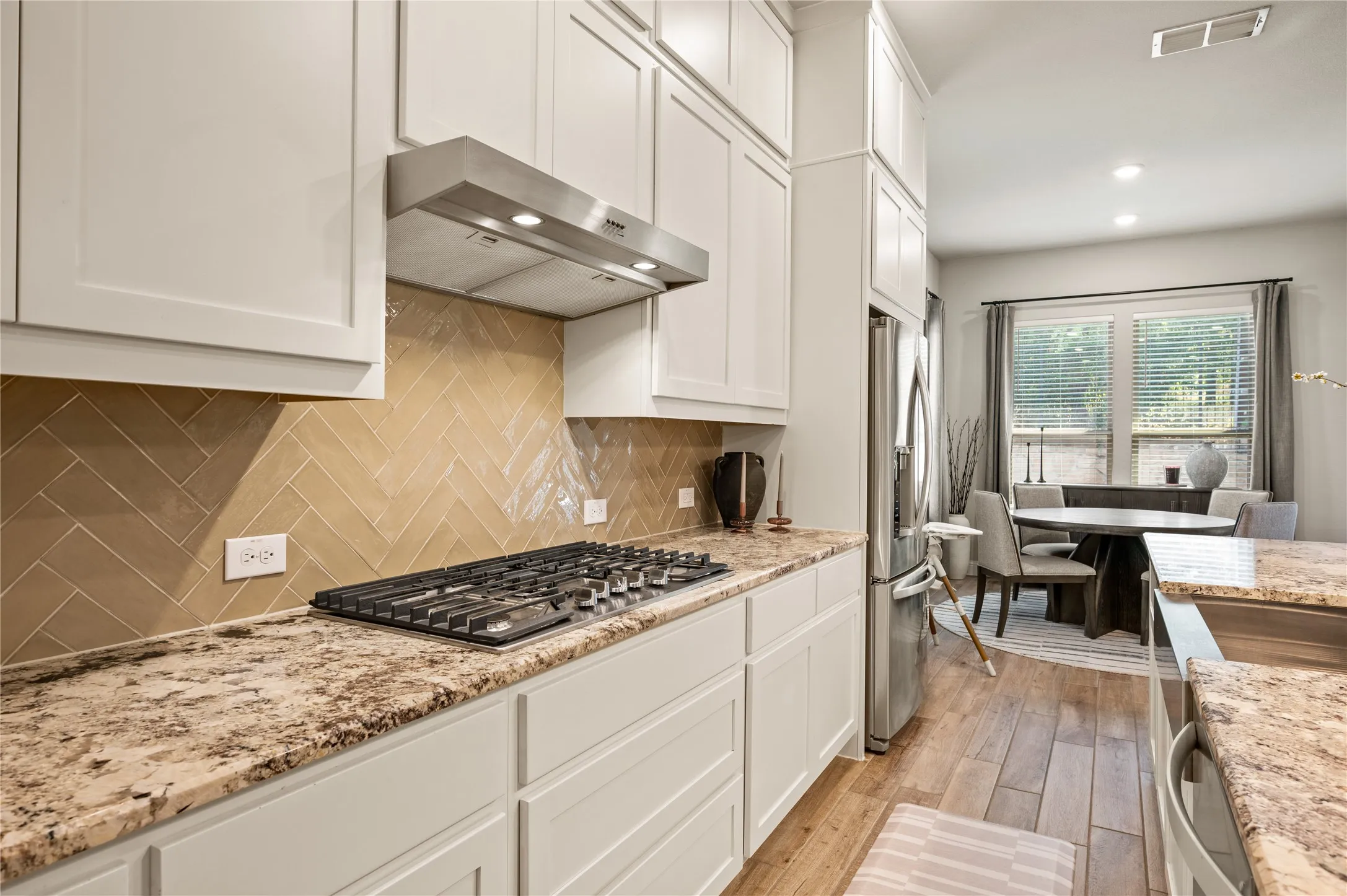 White shaker-style cabinetry, stainless steel range hood, herringbone tile backsplash, integrated gas cooktop, and wood-finish flooring