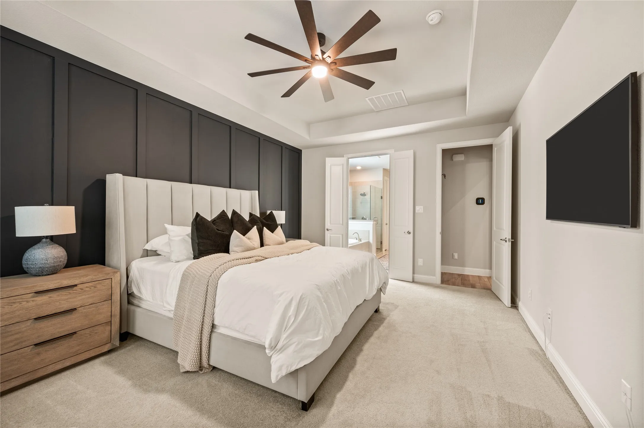 Accent wall with vertical paneling, ceiling fan with wood-finish blades, tray ceiling, wall-mounted television, and carpeted flooring