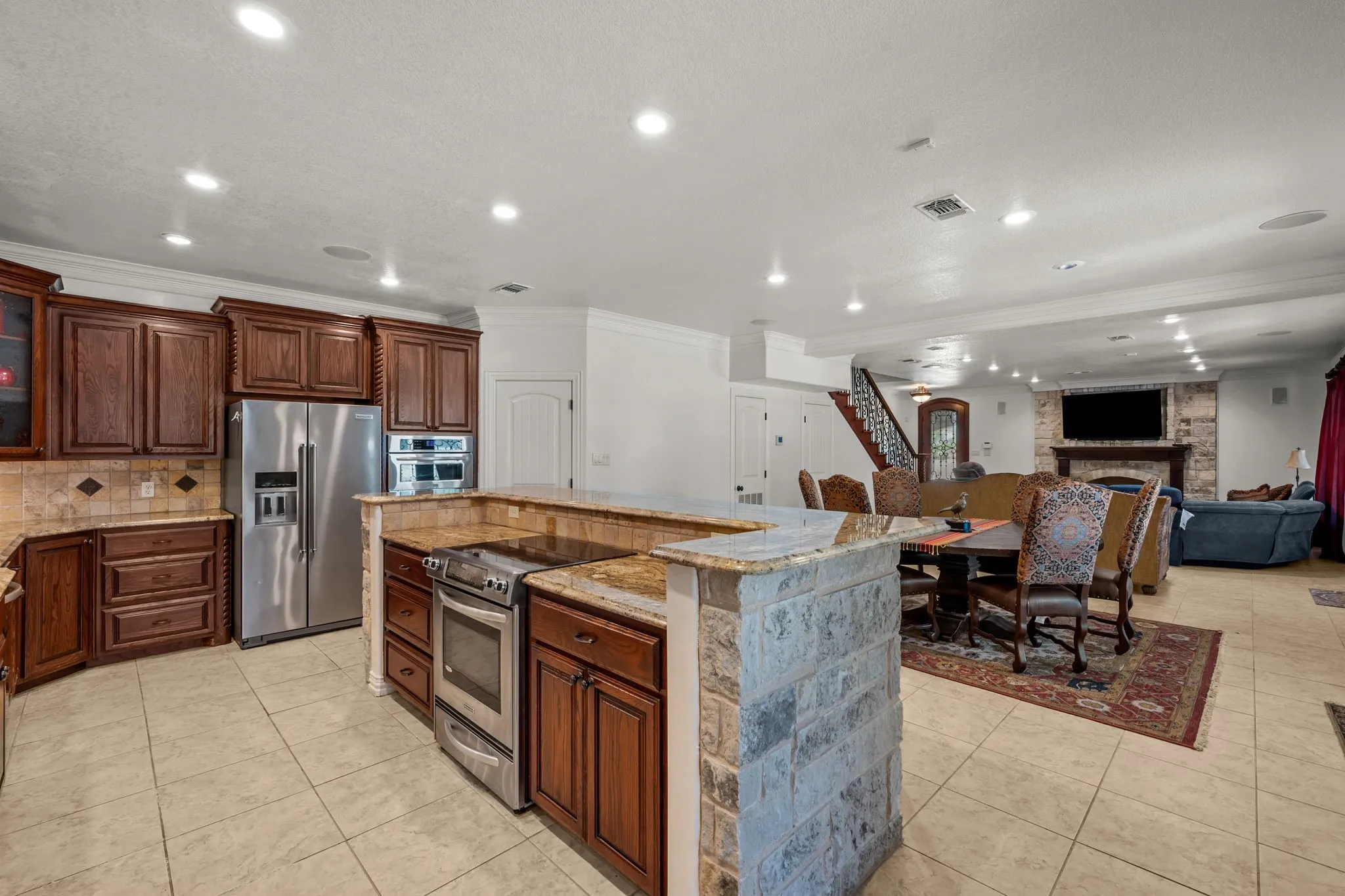 Open-concept living area with tile flooring, recessed lighting, a stone-clad kitchen island with counter seating, wood cabinetry, and a stone-clad fireplace