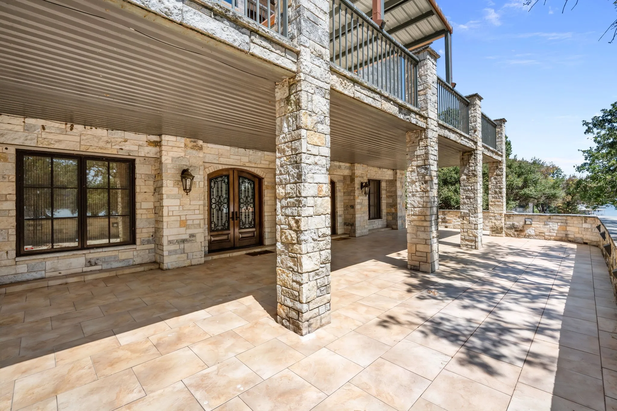 Expansive stone-paved patio with stone columns and walls, featuring a covered area with a corrugated metal ceiling, and ornate double entry doors