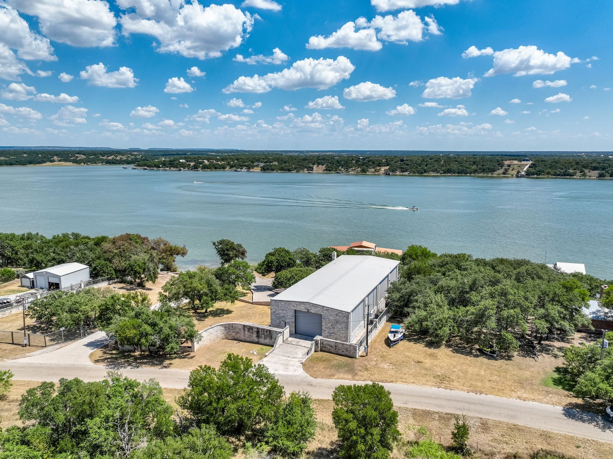 Waterfront property featuring a large stone and metal structure with a roll-up door, a separate metal building, mature trees, and a private driveway