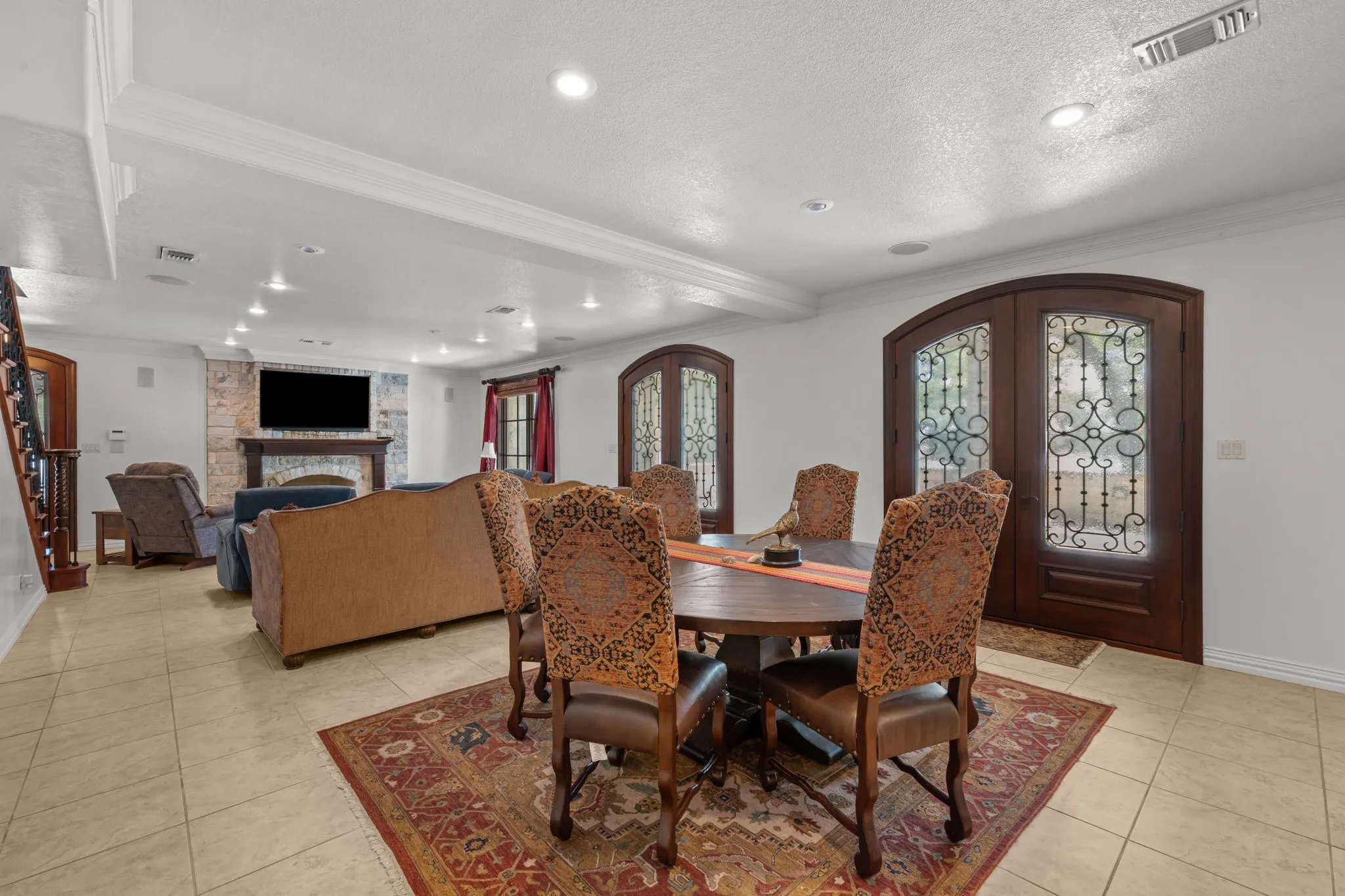 Tile flooring throughout, recessed lighting, stone fireplace surround with wood mantel, wood-finish staircase with wrought iron balusters, and arched wood-finish double doors with decorative glass inserts
