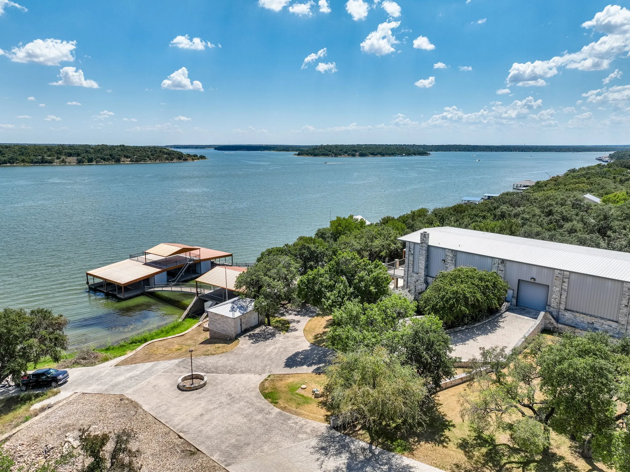 Waterfront property featuring a private boat dock with multiple slips, a large stone and metal-sided structure, a circular drive, and lush surrounding trees