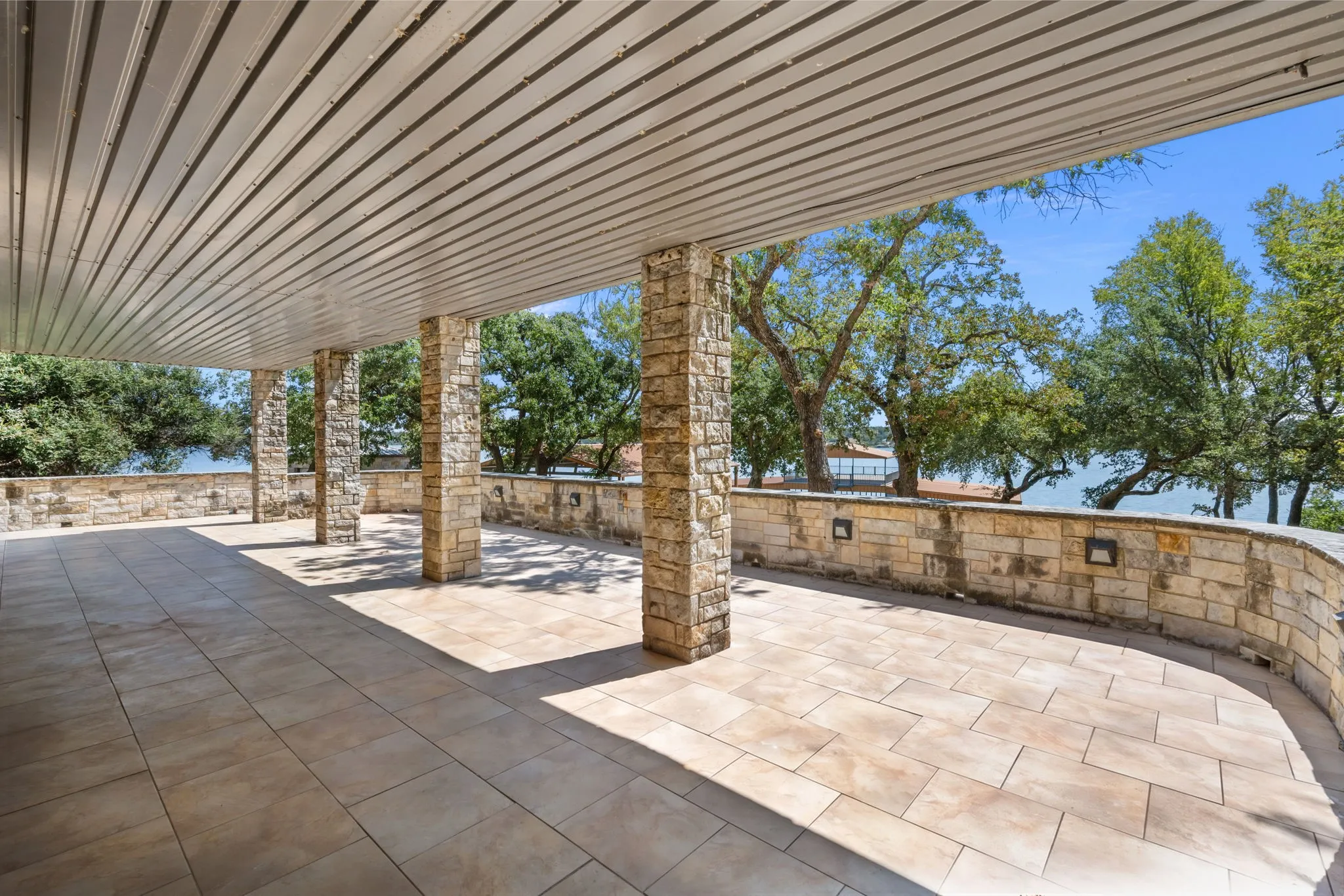 Covered outdoor patio featuring stone columns, stone knee wall, recessed wall lighting, a slatted ceiling, and large format tile flooring