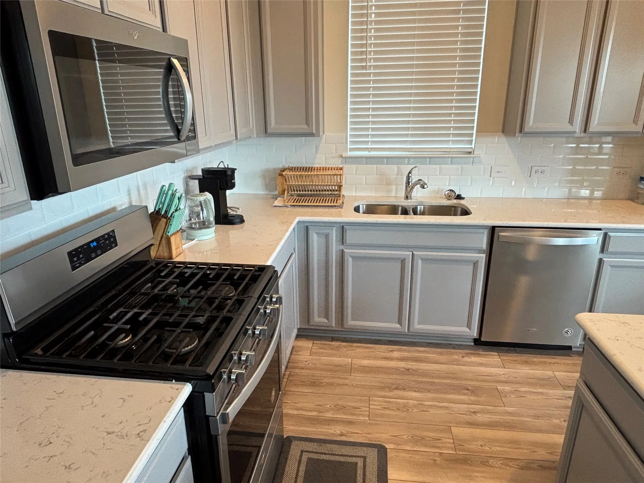Stainless steel appliances, light gray cabinetry, light-toned countertops, white subway tile backsplash, and wood-finish flooring