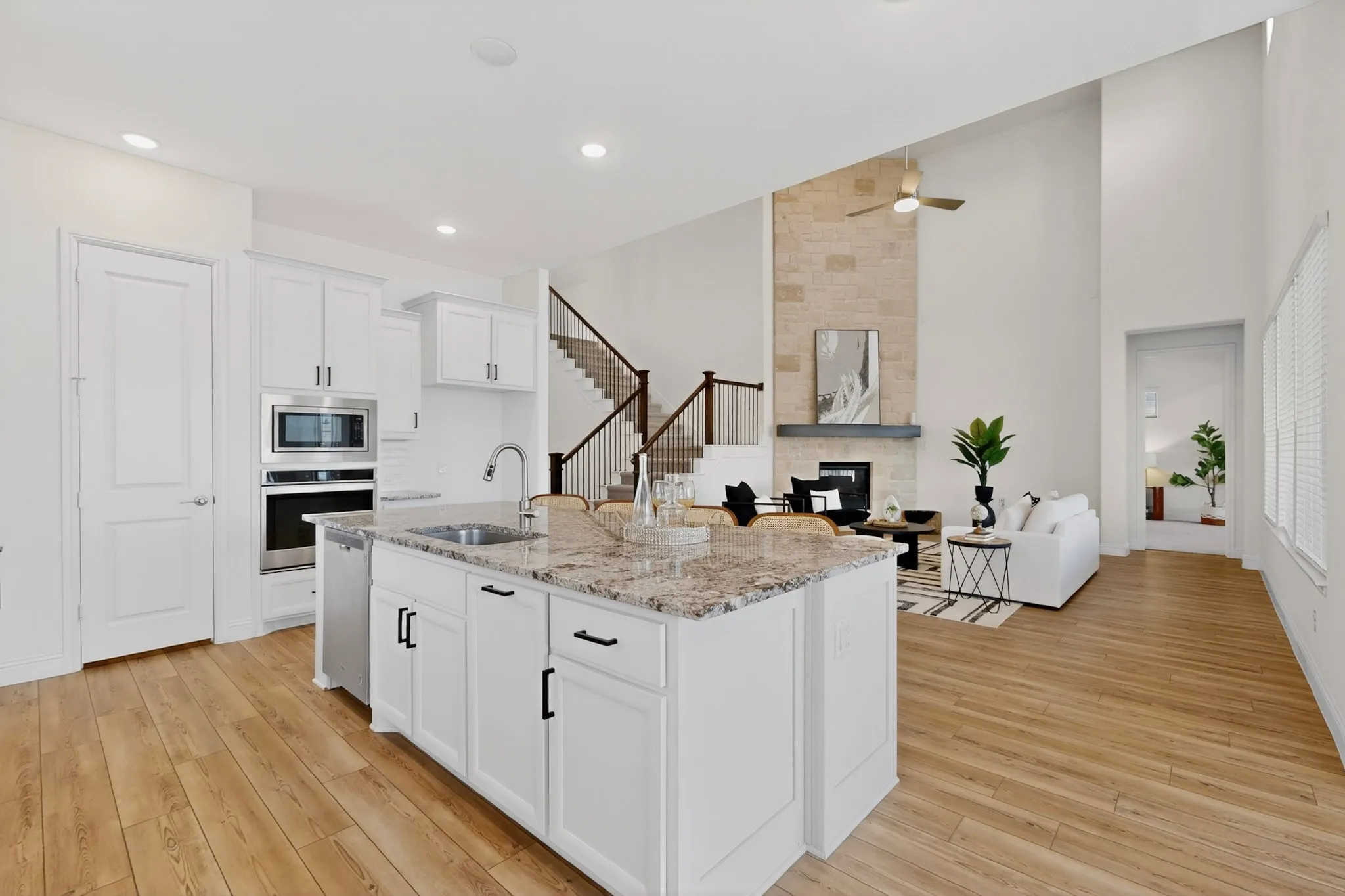 Open-concept living space featuring wood-finish flooring, a kitchen island with stone countertop, stainless steel appliances, a stone fireplace with mantel, and a staircase with metal balusters