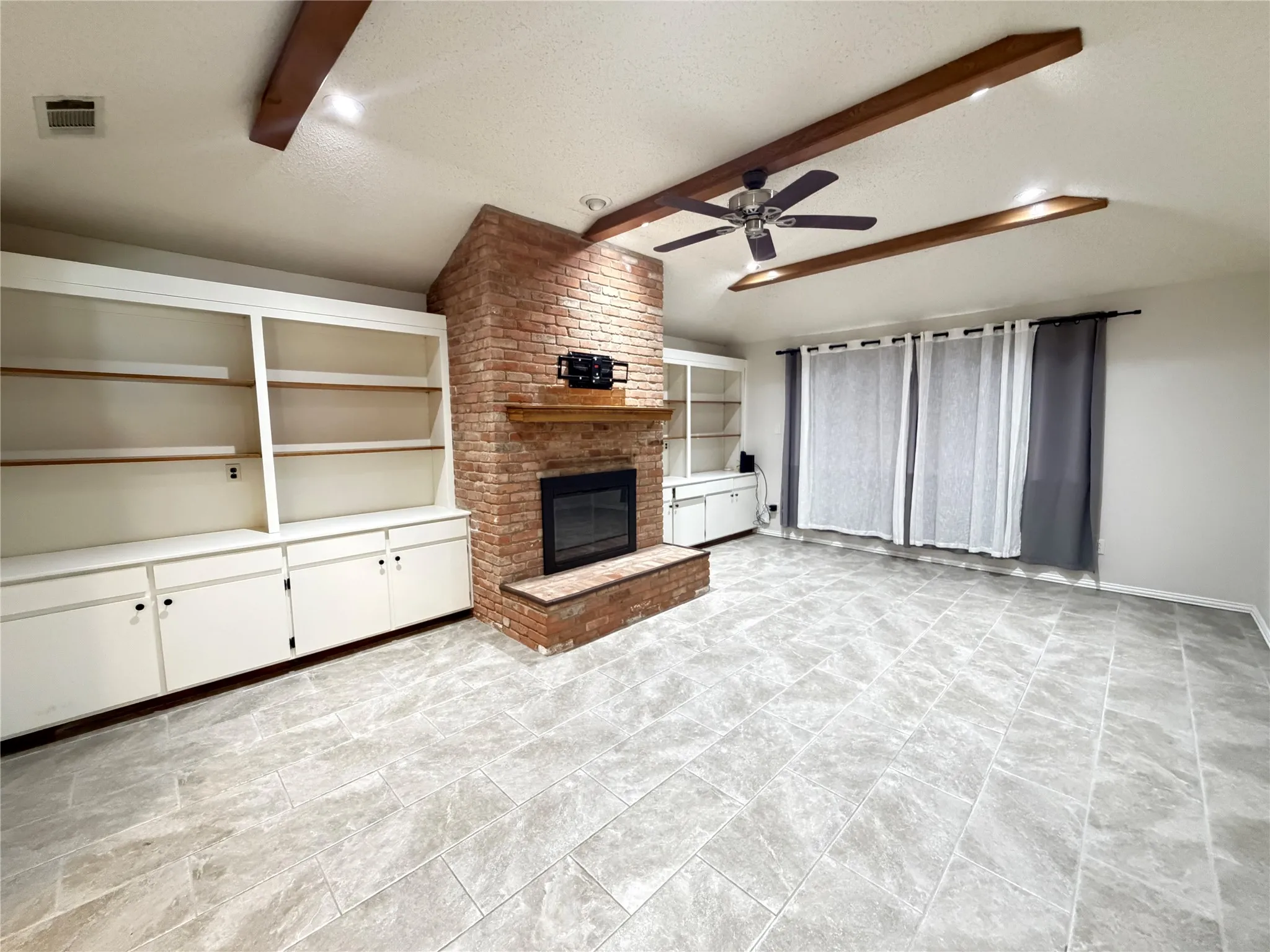 Brick fireplace with wood mantel, built-in shelving, wood-finish ceiling beams, ceiling fan, and large format tile flooring