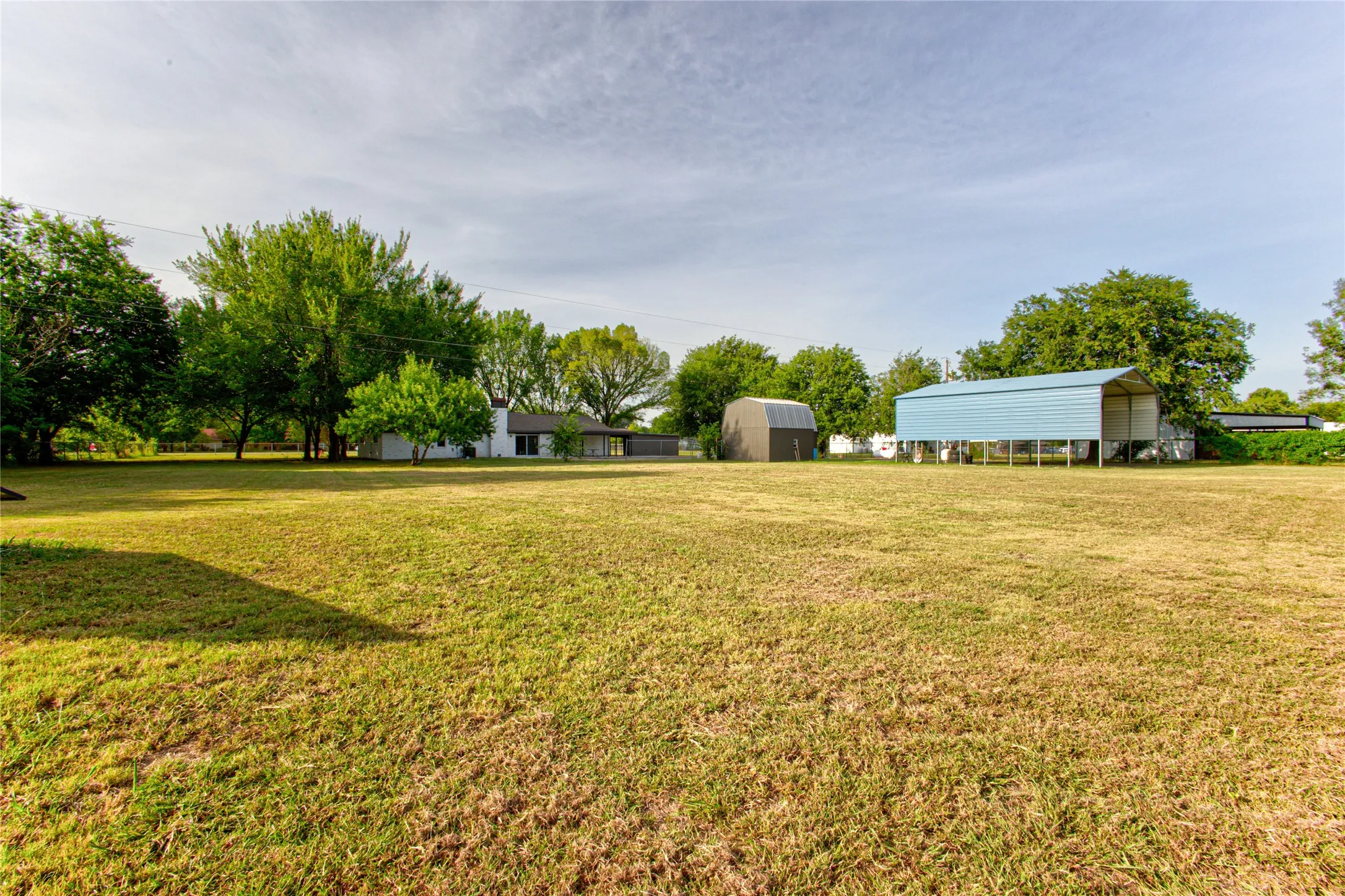 Expansive grassy yard with mature trees, a detached storage building, and an open-sided blue metal structure
