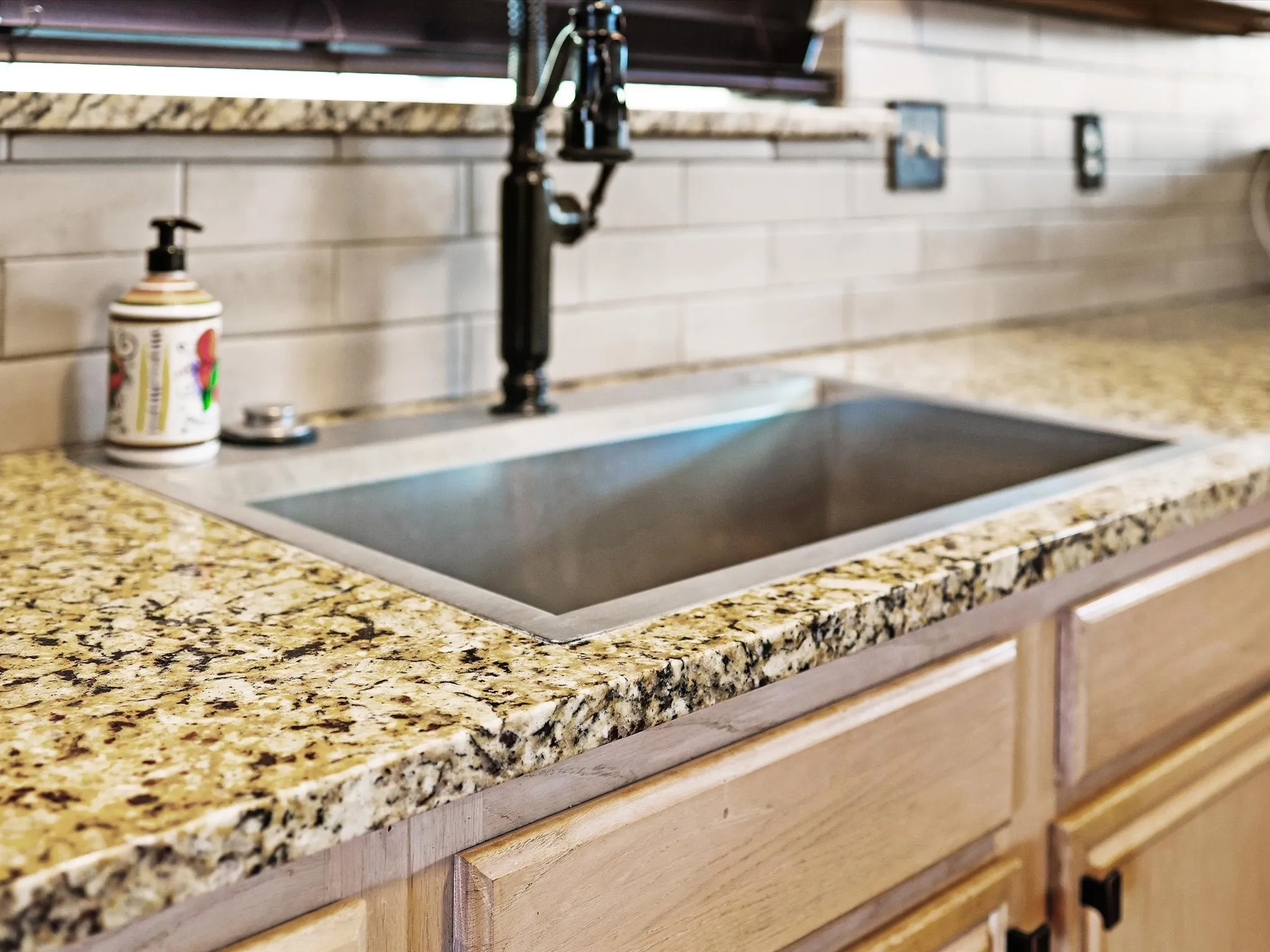 Granite countertops, undermount stainless steel sink, dark bronze gooseneck faucet, light wood-finish cabinetry, and subway tile backsplash