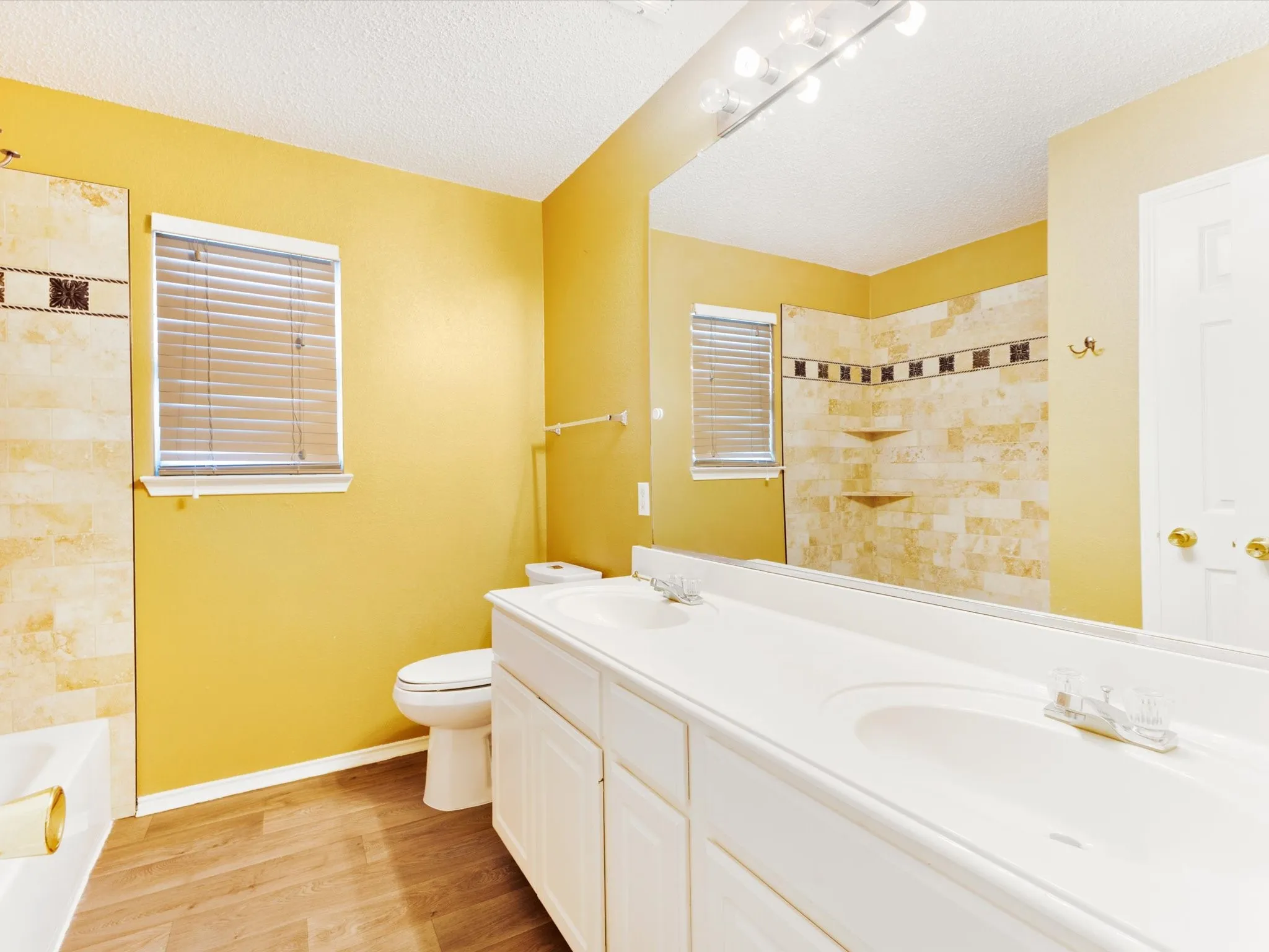 Double vanity with integrated sinks, wood-finish flooring, tiled shower surround, built-in shower shelving, and a window with blinds