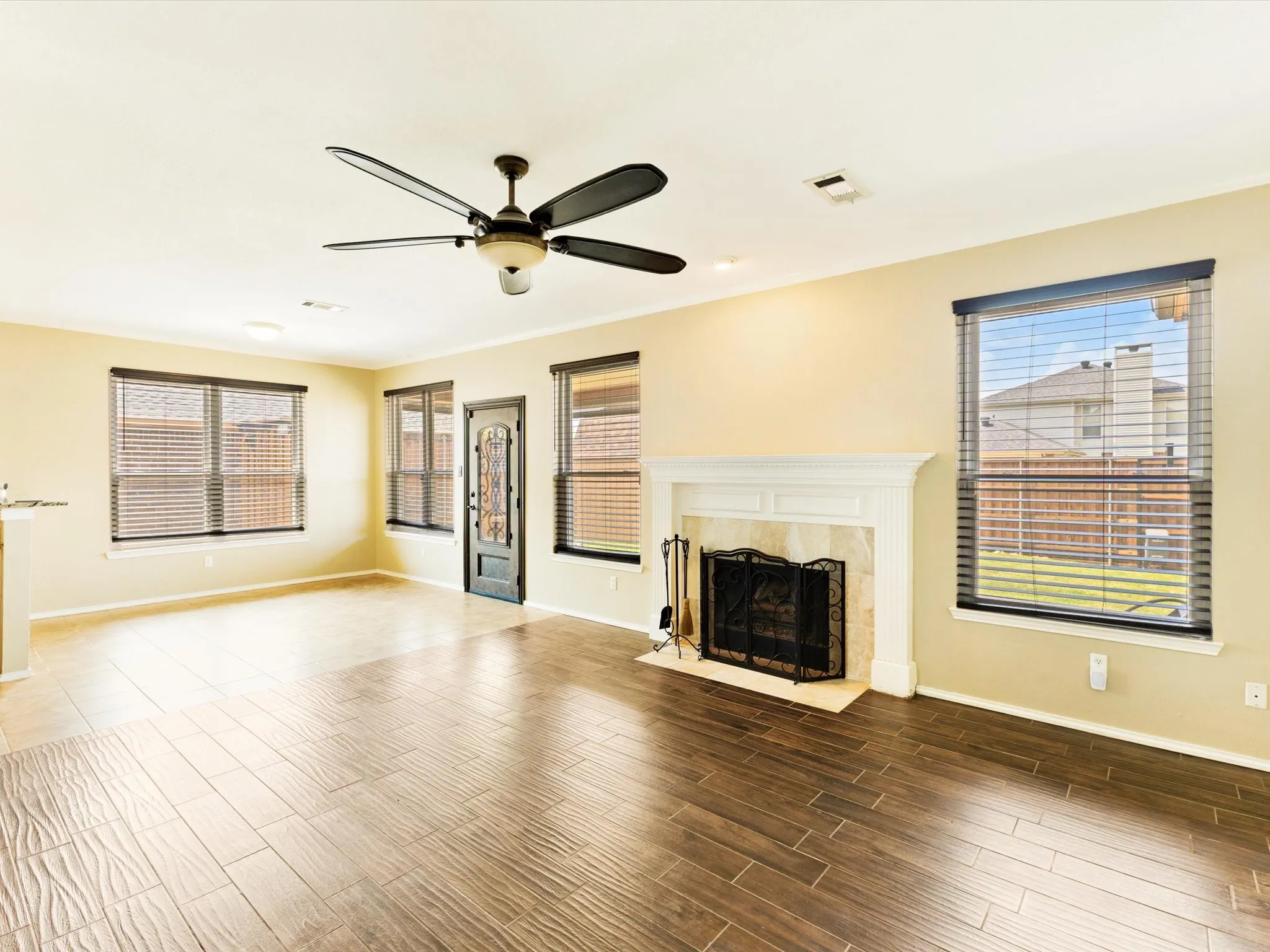 Wood-finish flooring, ceiling fan with integrated lighting, multiple windows with blinds, front entry door with decorative glass, and a fireplace with a white mantel and stone surround