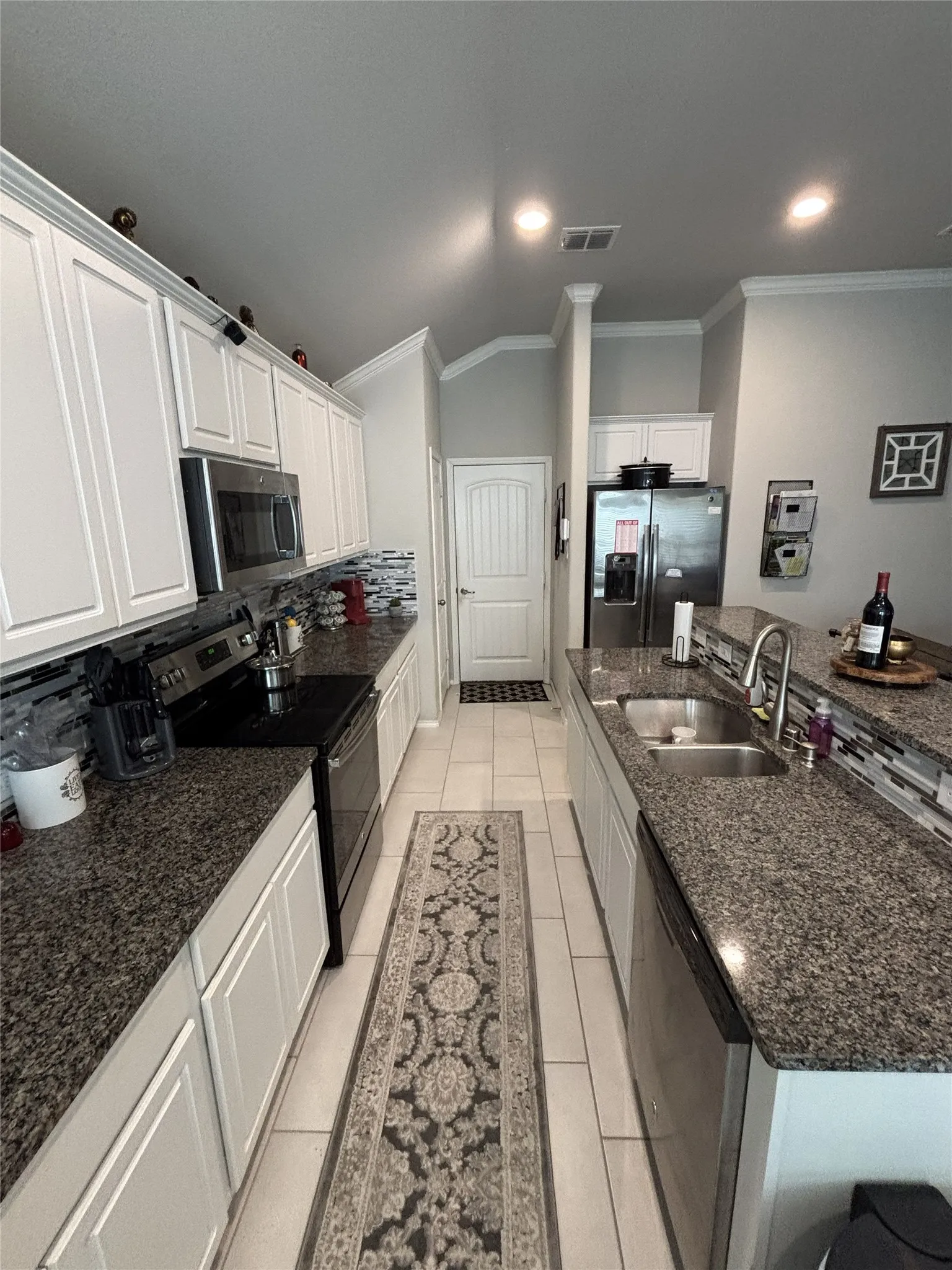 Galley kitchen featuring white cabinetry, granite countertops, tile flooring, stainless steel appliances, and a mosaic tile backsplash