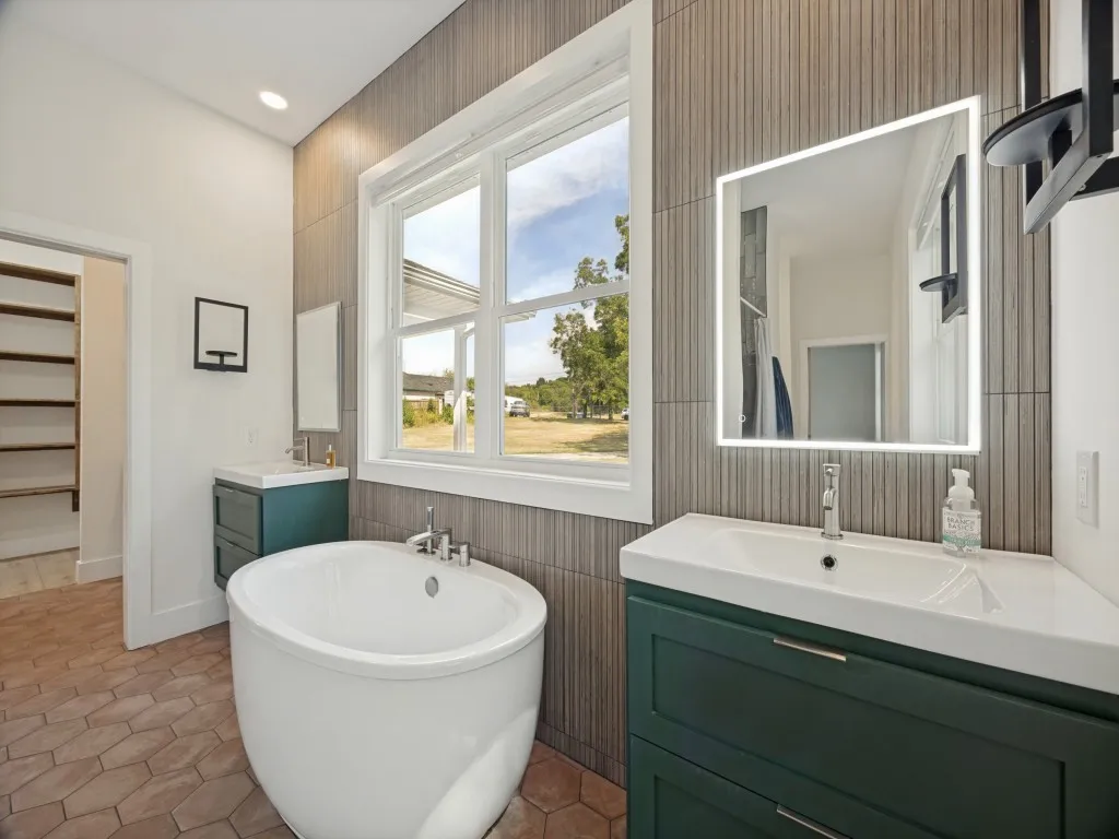 Freestanding oval soaking tub, dual vanities with integrated sinks, illuminated mirrors, and patterned wall tiling; opens to the laundry room
