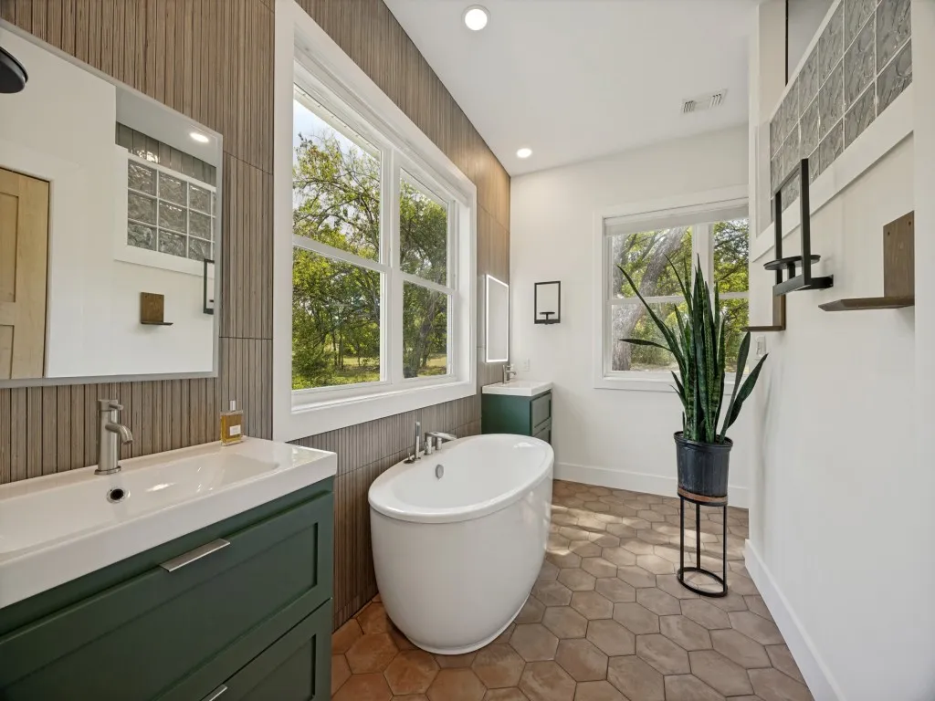 Freestanding soaking tub, dual vanity with green cabinetry, large windows, recessed lighting, and hexagonal floor tiling