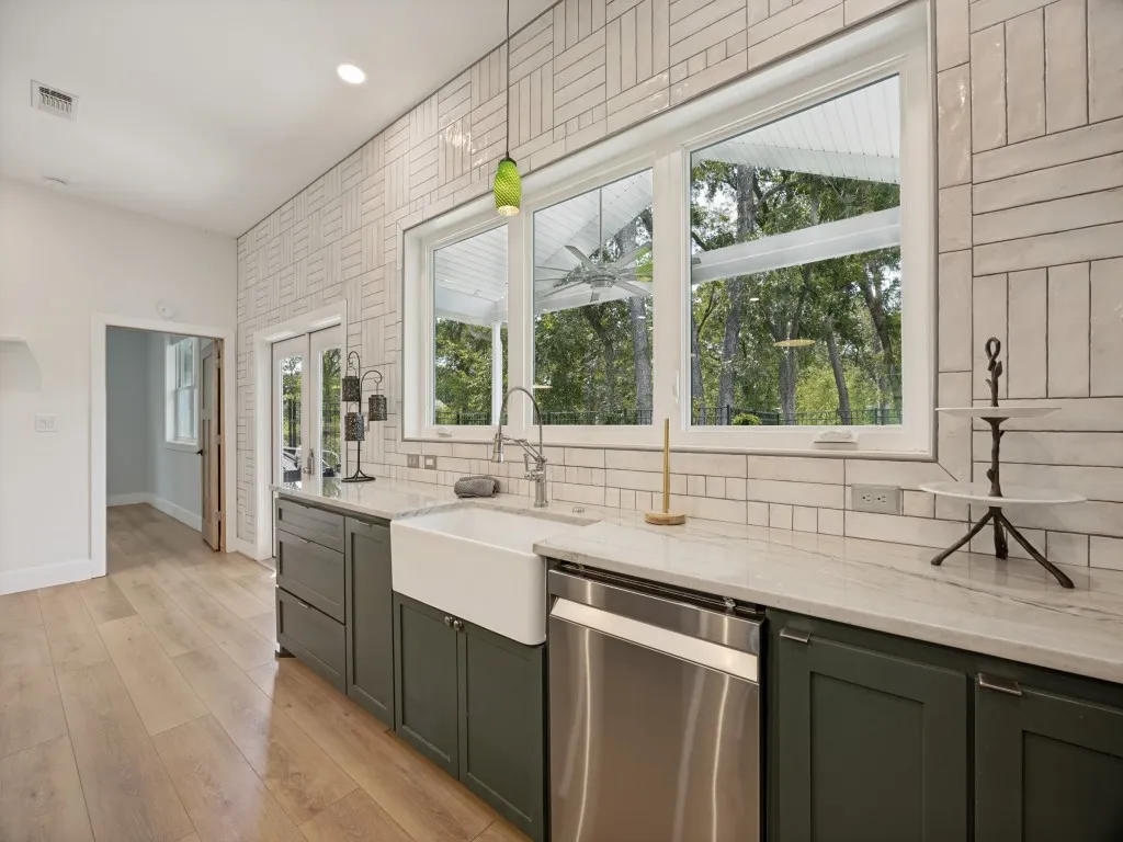 Fully updated kitchen with wood-finish flooring, white subway tile backsplash, white farmhouse sink, stainless steel dishwasher, and green cabinetry