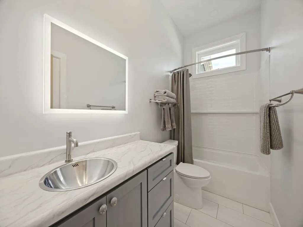 Modern bathroom featuring a backlit LED mirror, floating vanity with dark cabinetry and white stone countertop, stainless steel vessel sink, ceramic tile flooring, and a walk-in shower with subway tile surround and glass enclosure
