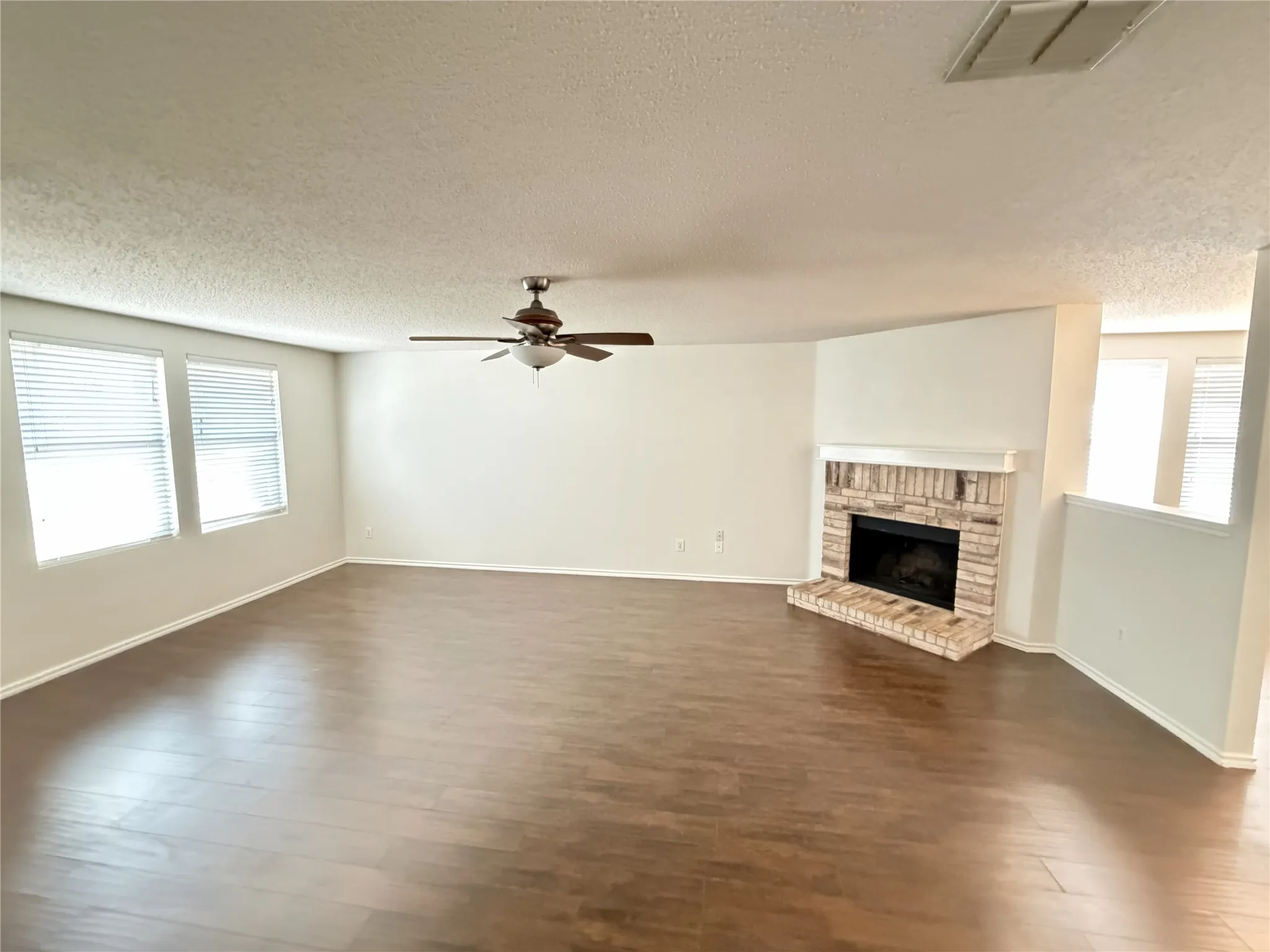 Spacious room featuring wood-finish flooring, multiple windows with blinds, a ceiling fan, and a corner fireplace with a stone surround