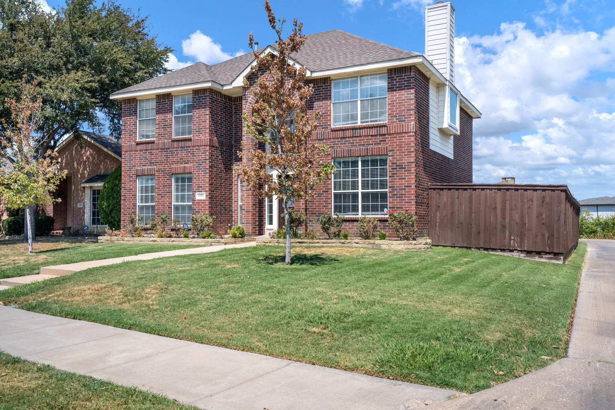 Brick exterior with a prominent chimney, large windows, established landscaping, and a privacy fence
